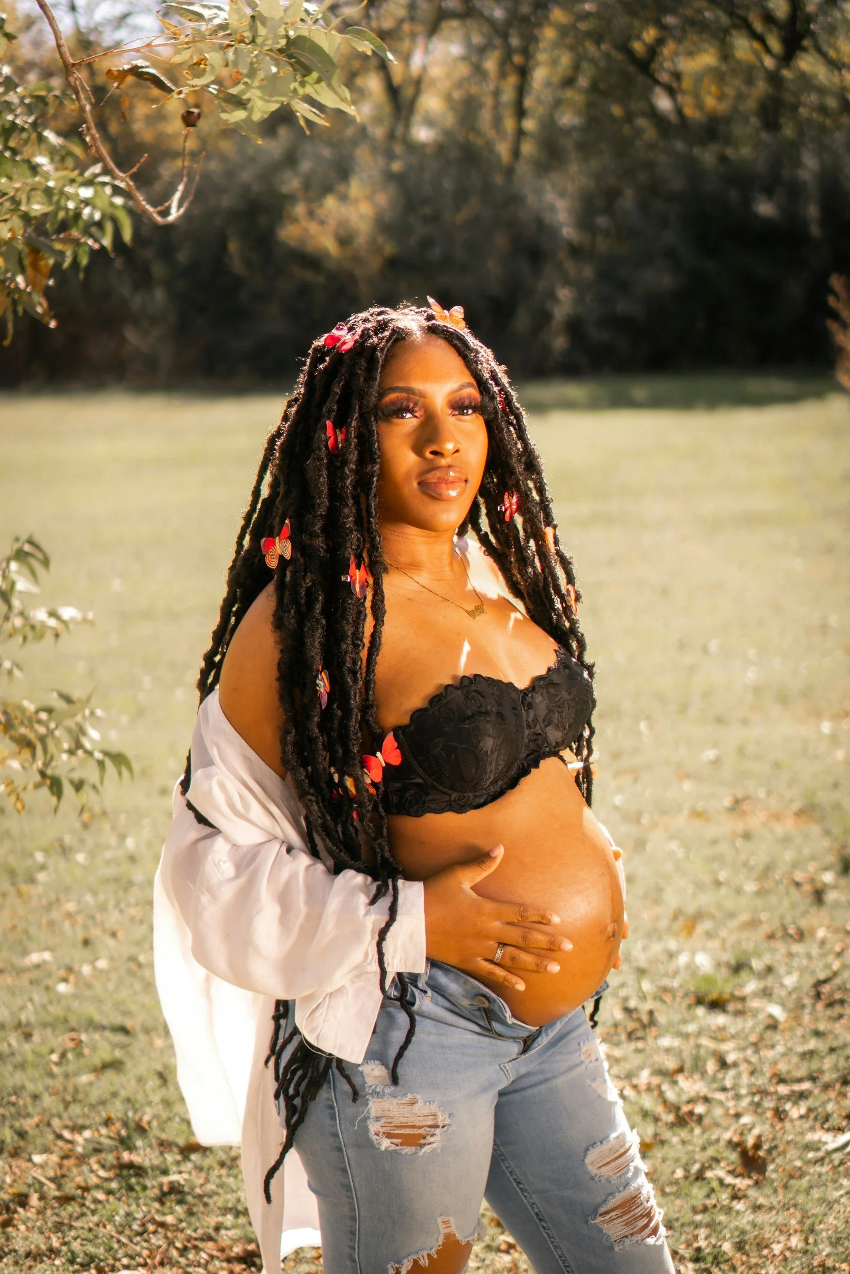 A pregnant woman standing outdoors near a tree, wearing a black bra, distressed jeans, and a white shirt off her shoulders, with colorful butterfly hair accessories in her long dreadlocks.