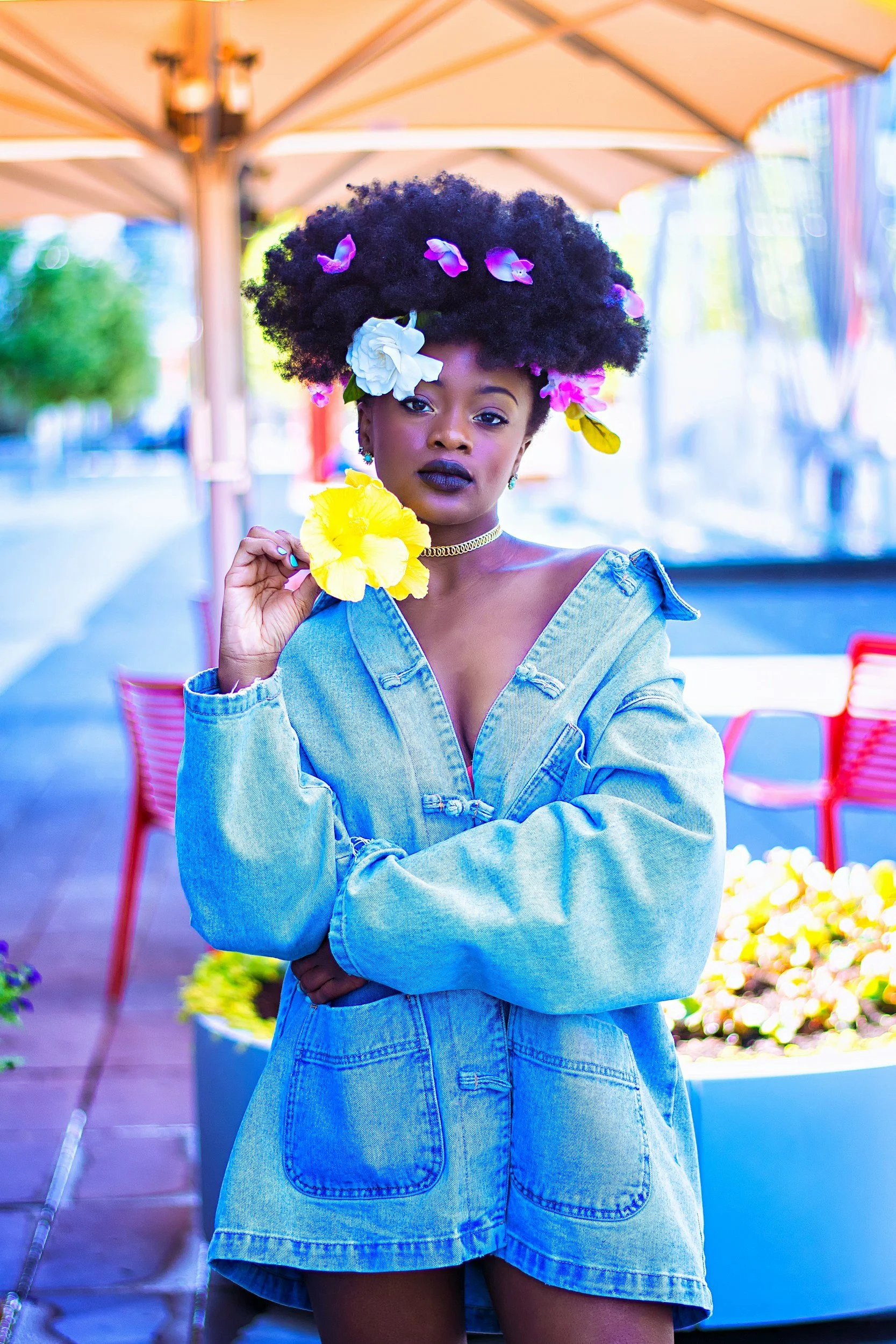 A woman with dark lipstick and bold makeup, wearing a denim dress and a gold choker necklace, is holding a yellow hibiscus flower near her face. She has an elaborate afro hairstyle decorated with pink and purple flowers. The background features an outdoor setting with large umbrellas, colorful chairs, and potted plants.