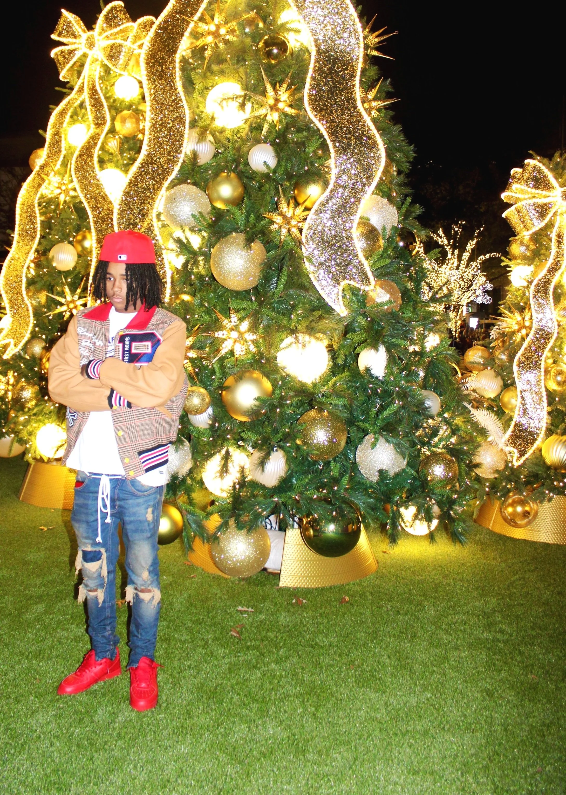 A young man standing with arms crossed in front of a large, decorated Christmas tree at night. The tree is adorned with gold, white, and glittery ornaments and ribbon with bright lights.