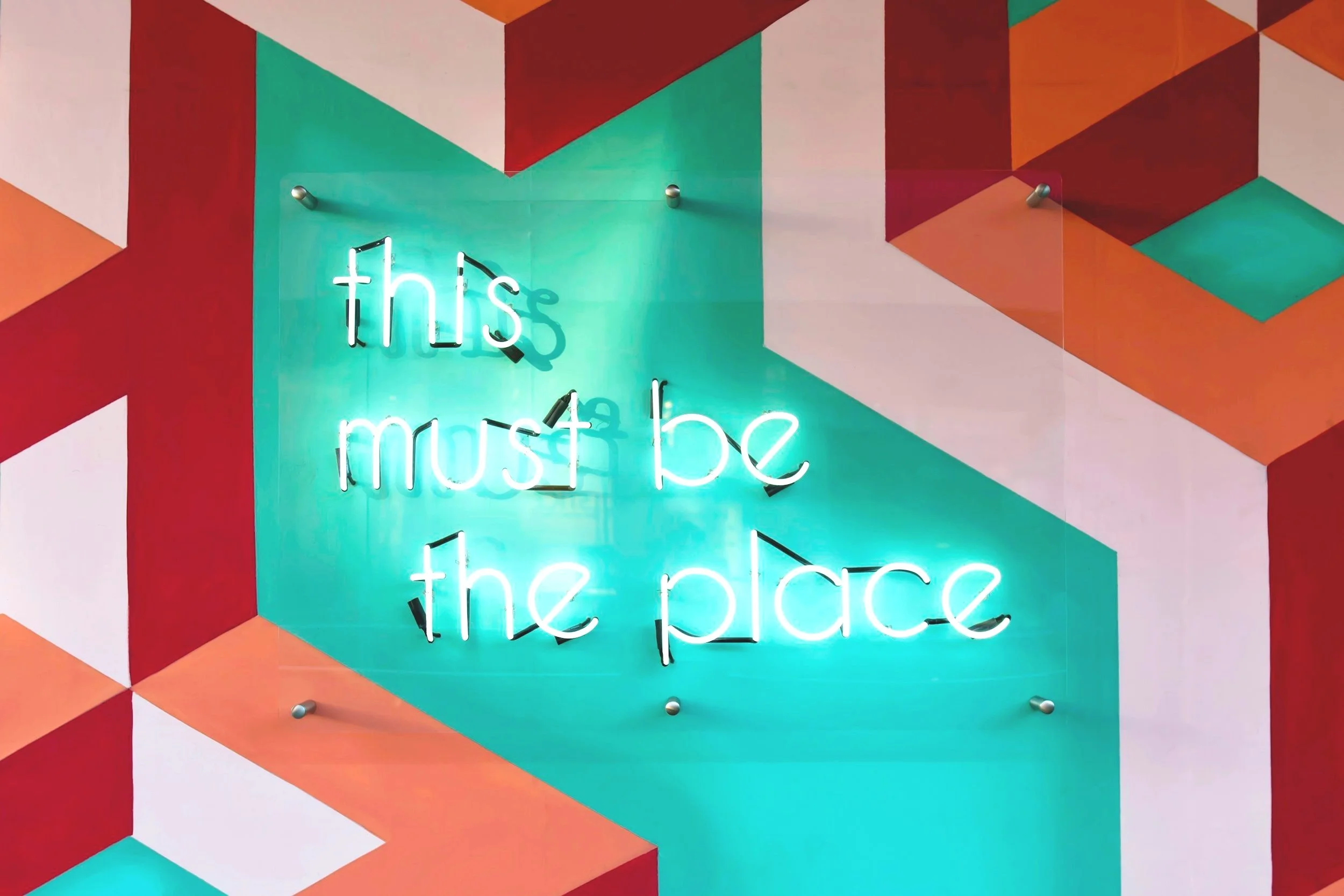 Colorful geometric wall with a neon sign that reads 'this must be the place' in cursive font.