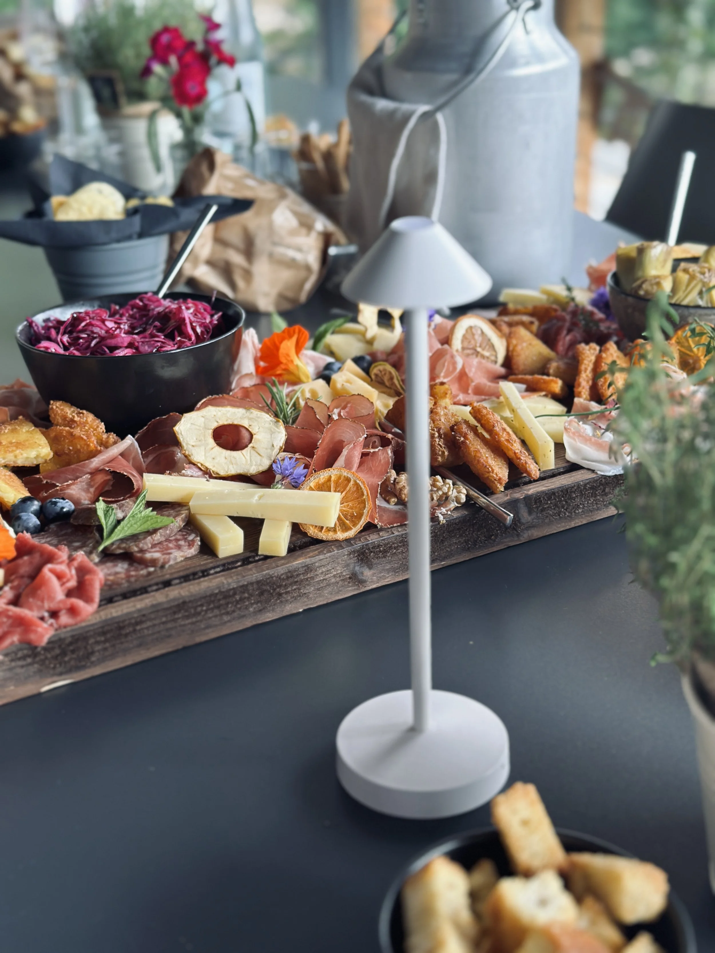 Tabella con un vassoio di antipasti, tra cui affettati, formaggi, frutta secca e verdure, accompagnati da ciotole di insalata e pane. In primo piano, un vassoio di crocchette e un piatto di insalata rosso-viola con cetrioli. Sullo sfondo, elementi decorativi e un lampada da tavolo.