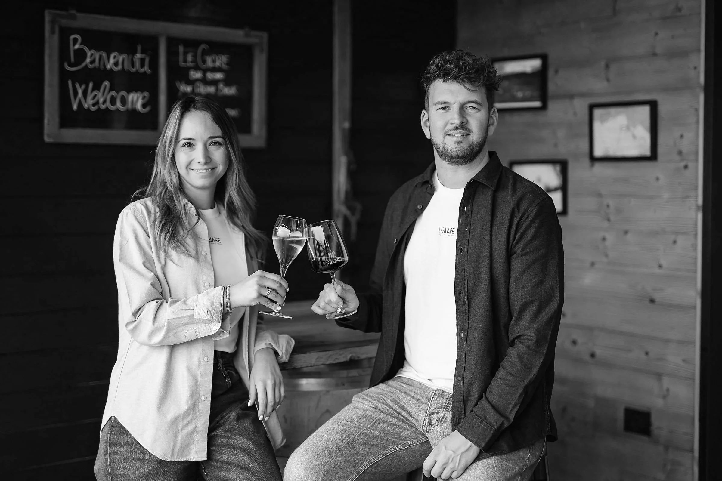 Due persone che sorridono e alzano bicchieri di vino in un ambiente interno con pareti di legno, un cartellone con scritto 'Benvenuti' e 'Welcome' in inglese e altre immagini alle pareti.