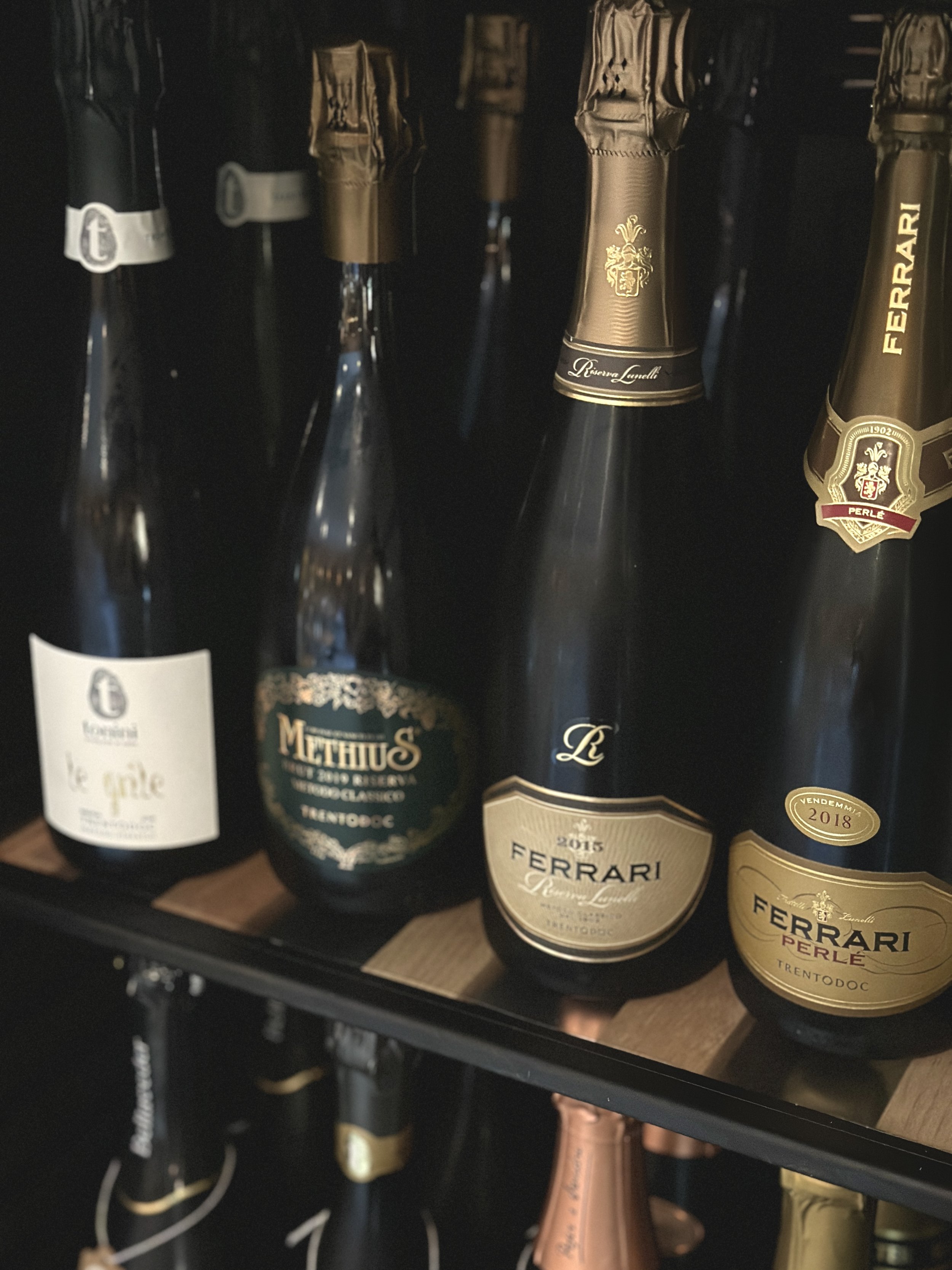 In questa immagine, ci sono diverse bottiglie di Trentodoc in uno scaffale di un frigo. Le bottiglie sono di marchi vari, con etichette dorate e nere, tra cui si evidenziano le bottiglie di Ferrari, alcune delle quali sono del 2015 e altre del 2018.