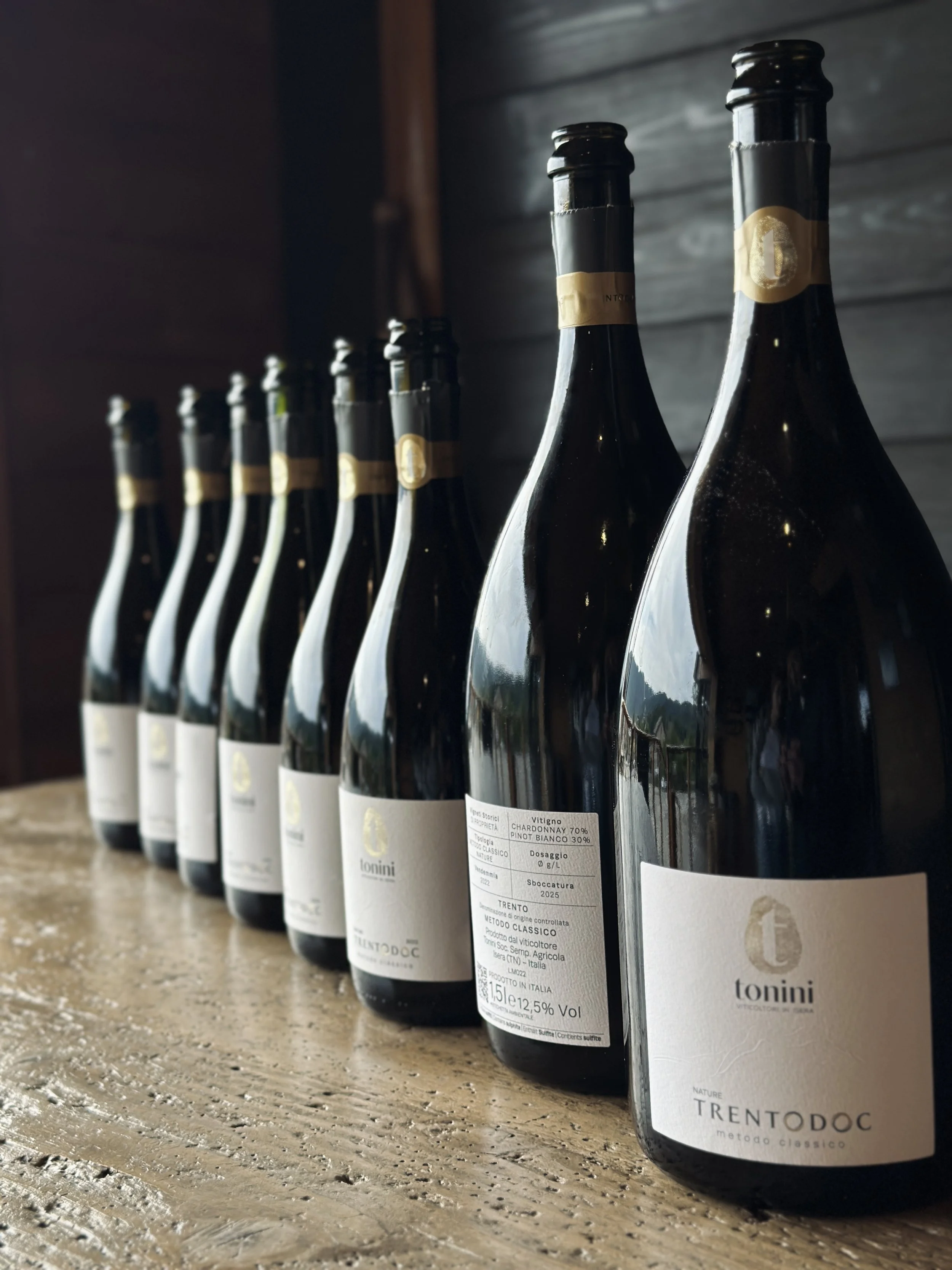 Sei bottiglie di vino spumante sulla superficie di legno, con etichette che indicano che si tratta di Trento DOC, metodo classico, Chardonnay 70% e Pinot Bianco 30%, annata 2022.