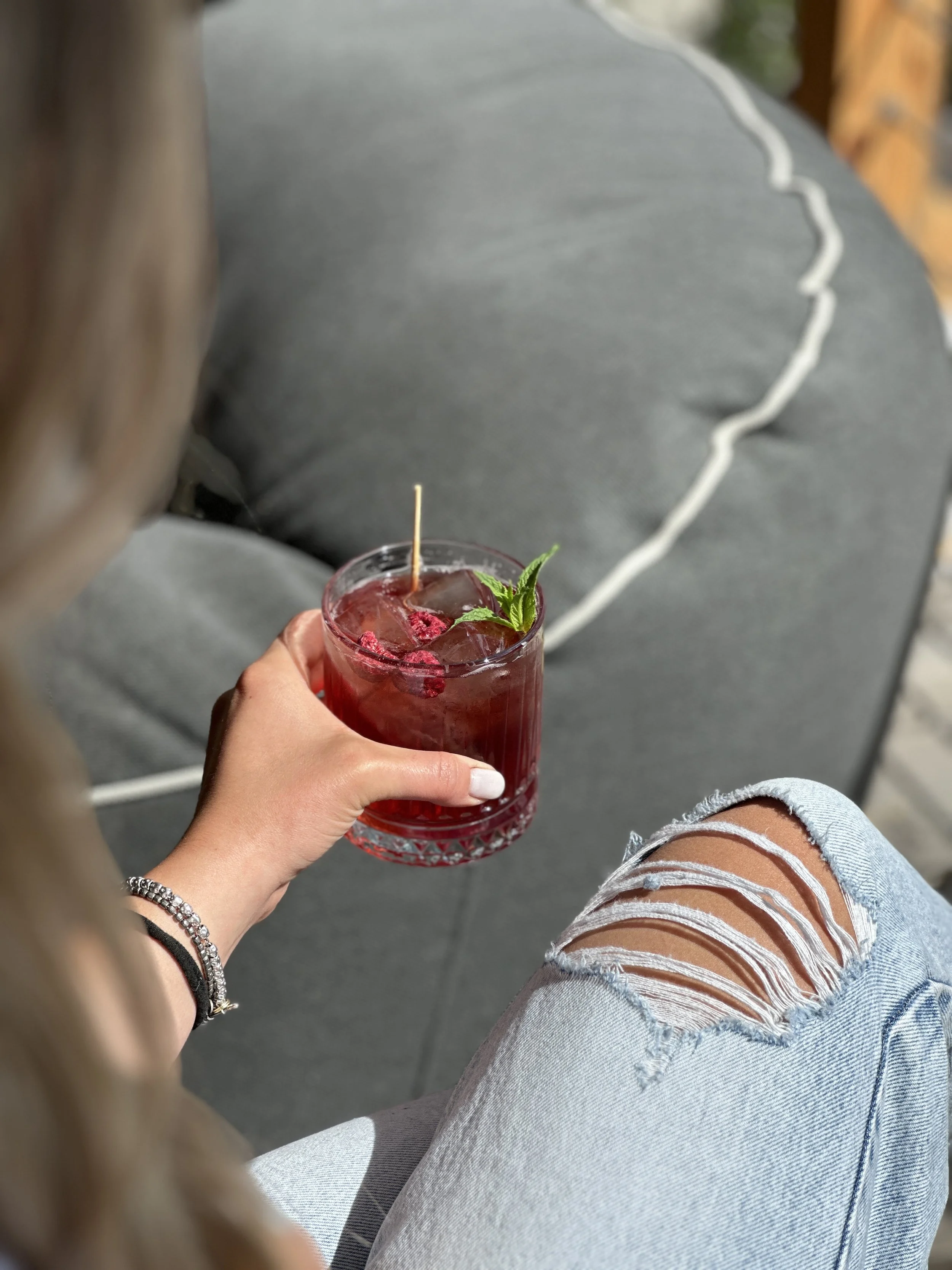 Una persona tiene in mano un bicchiere di cocktail rosso con ghiaccio e una foglia di menta, indossa braccialetti e jeans strappati, seduta su un pouff .