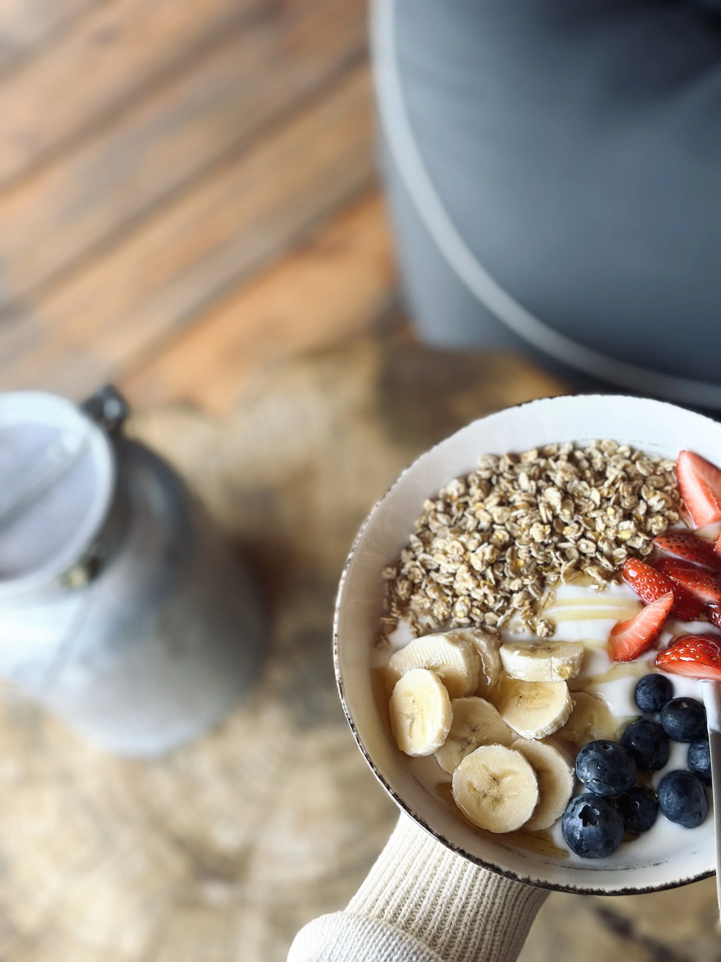 Una ciotola di yogurt decorata con fragole a fette, mirtilli,  banana a pezzetti, granola e miele, tenuta da una persona con un maglione a righe di colore beige.