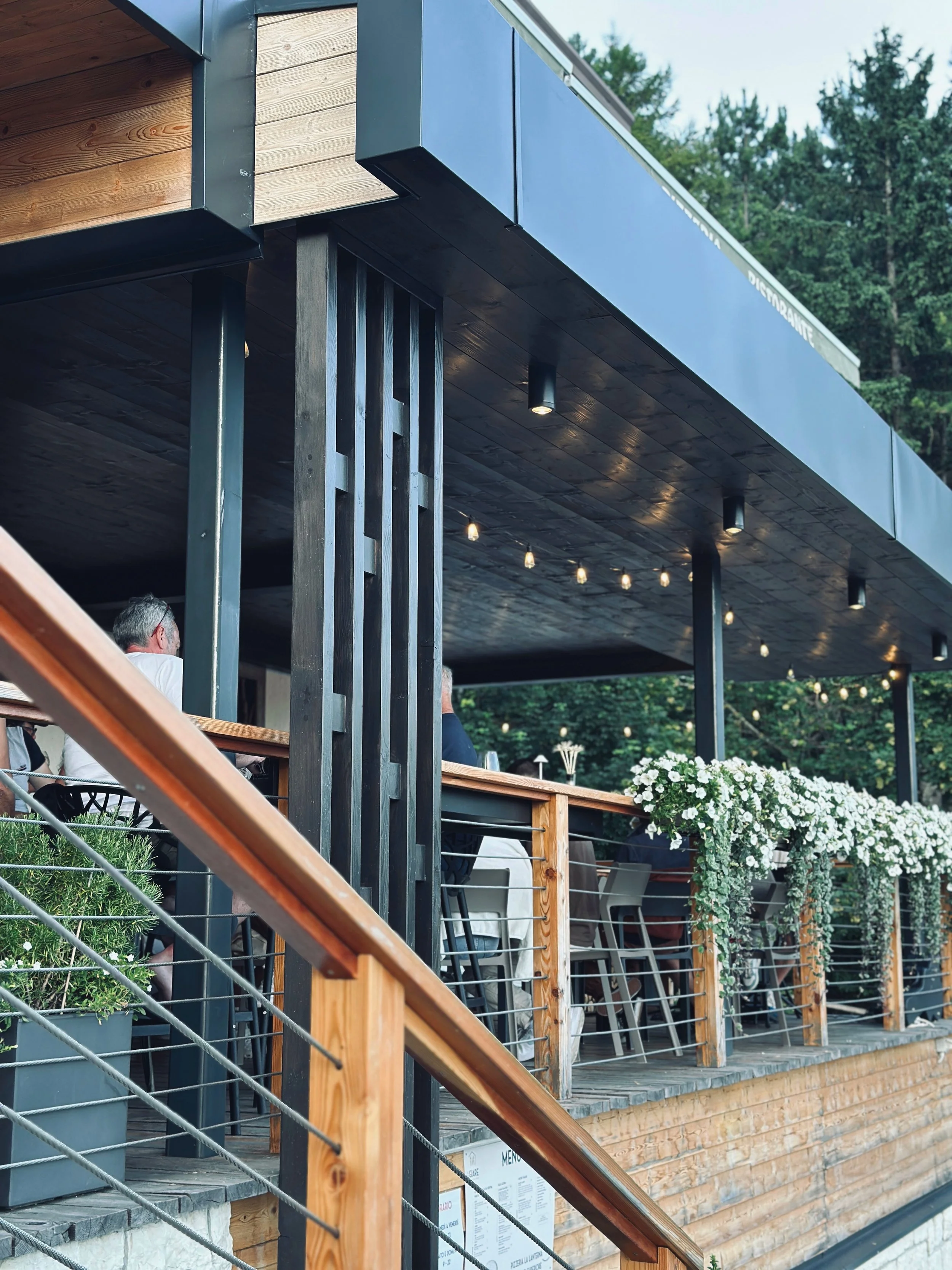 Vista di un ristorante o caffè all'aperto con tavoli e sedie, decorato con fiori bianchi, struttura in legno e illuminazione soffusa, circondato da alberi e natura.
