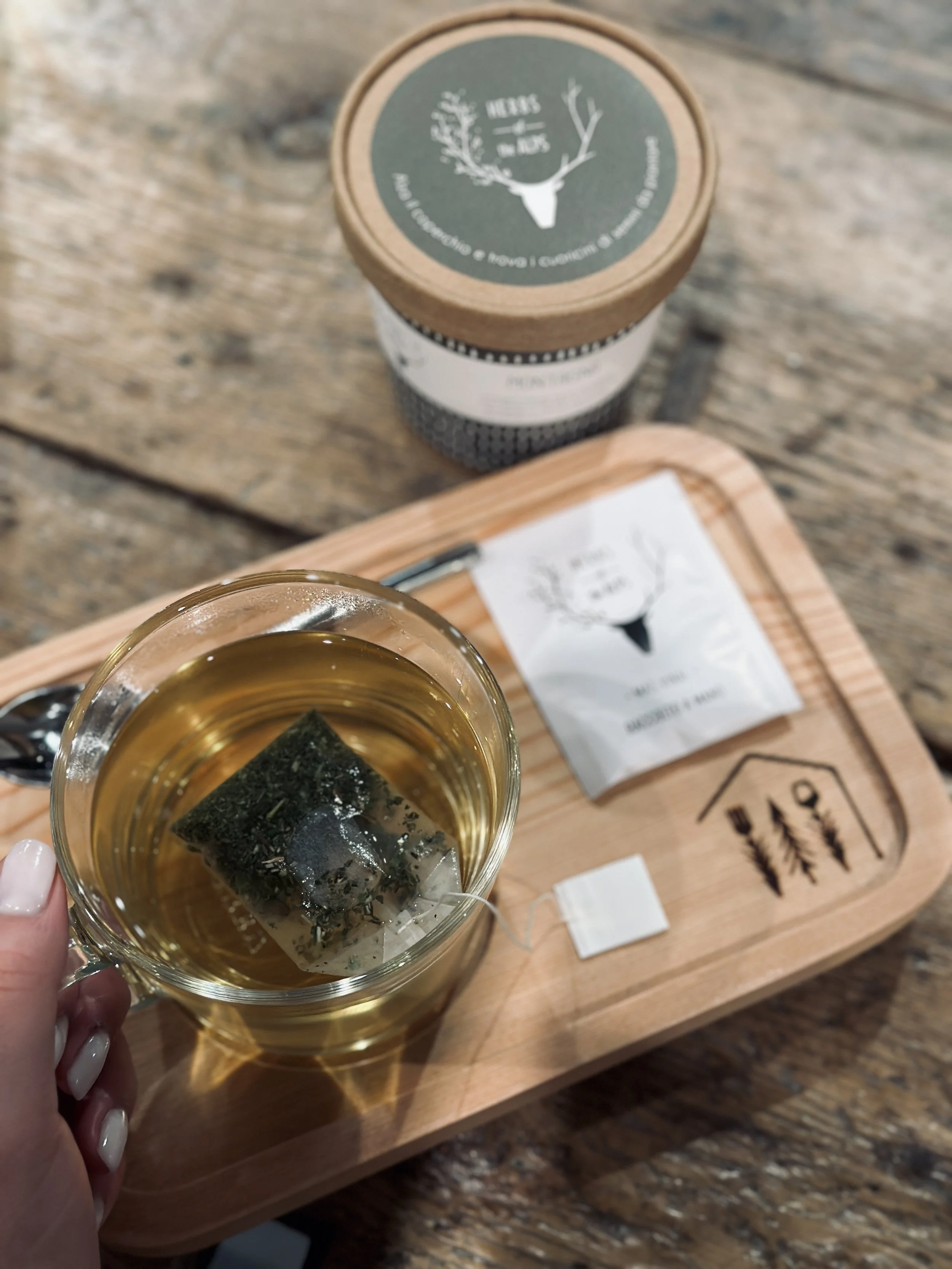 Bicchierino di tè con foglie di tè in infusione, sopra un vassoio di legno con un menu e una scatola decorativa con alci e alberi, su un tavolo di legno rustico.
