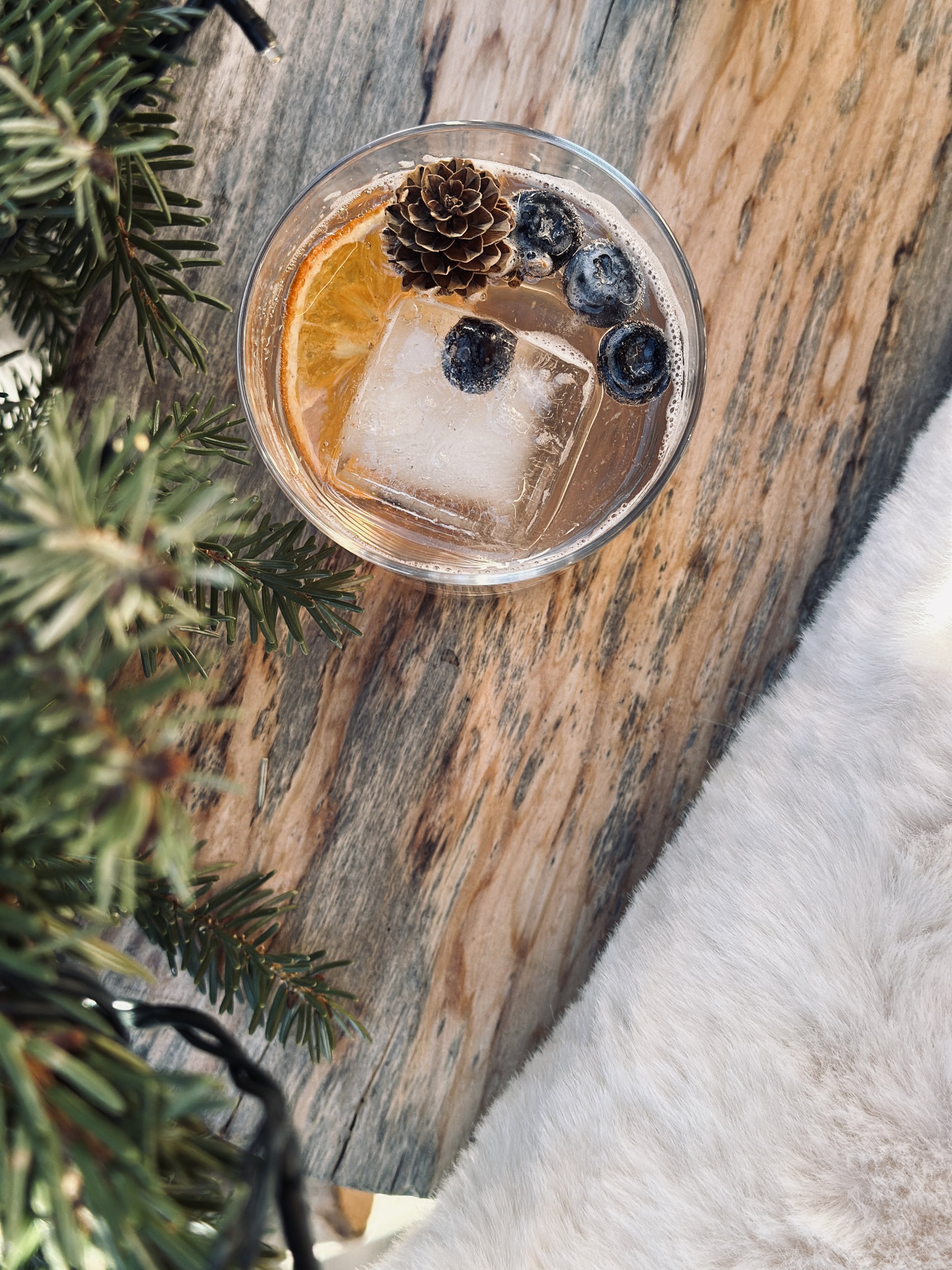 Bicchiere trasparente con cocktail, fetta di arancia, mirtilli, un pigne, ghiaccio, posizionato su un tavolo di legno, con rametto di pino a sinistra e una superficie bianca a destra.