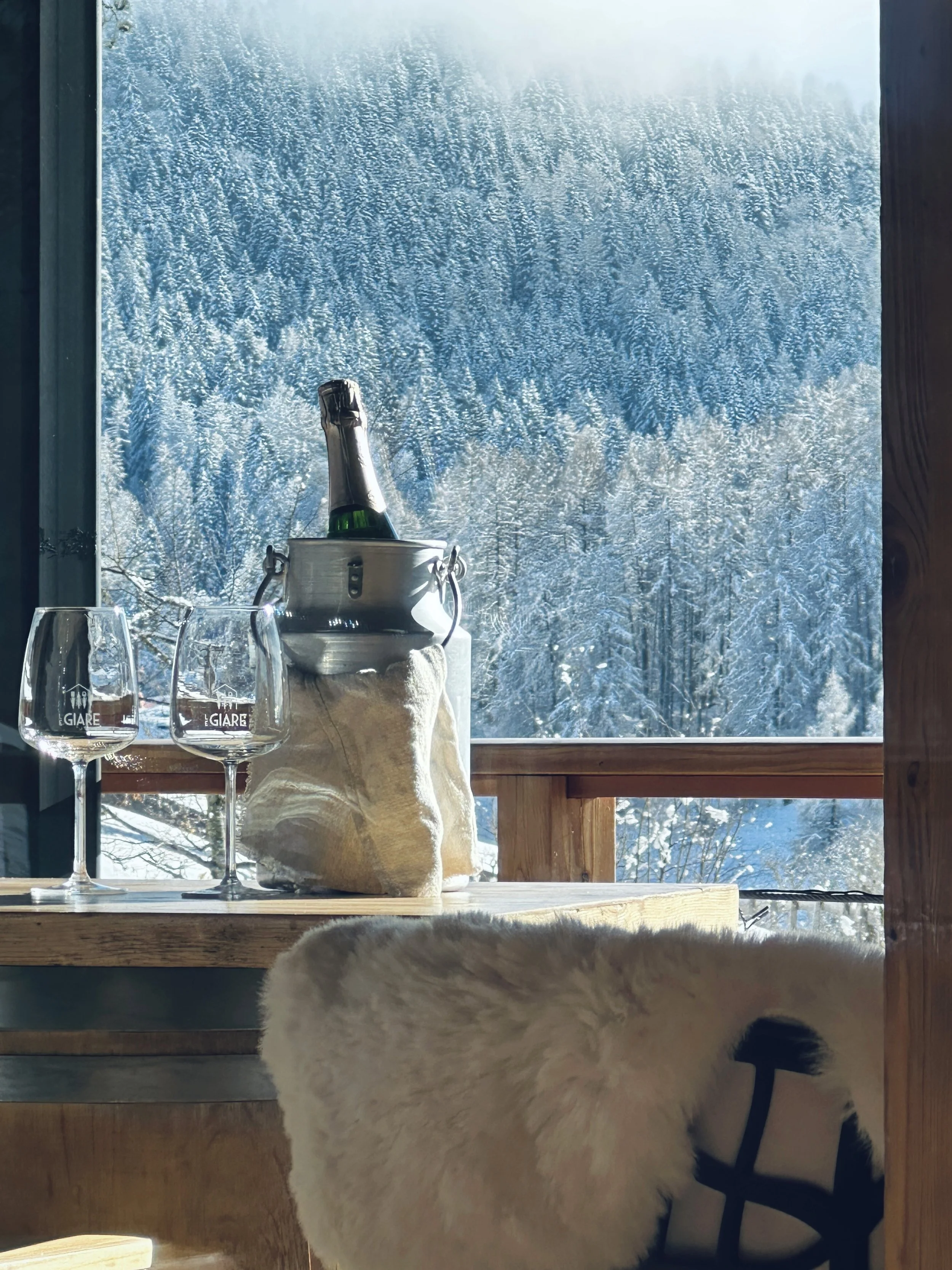Interno di una baita con vista sulla foresta innevata attraverso una finestra, con bottiglia di champagne, due bicchieri e una coperta sul tavolo, e un cane sulla sedia.