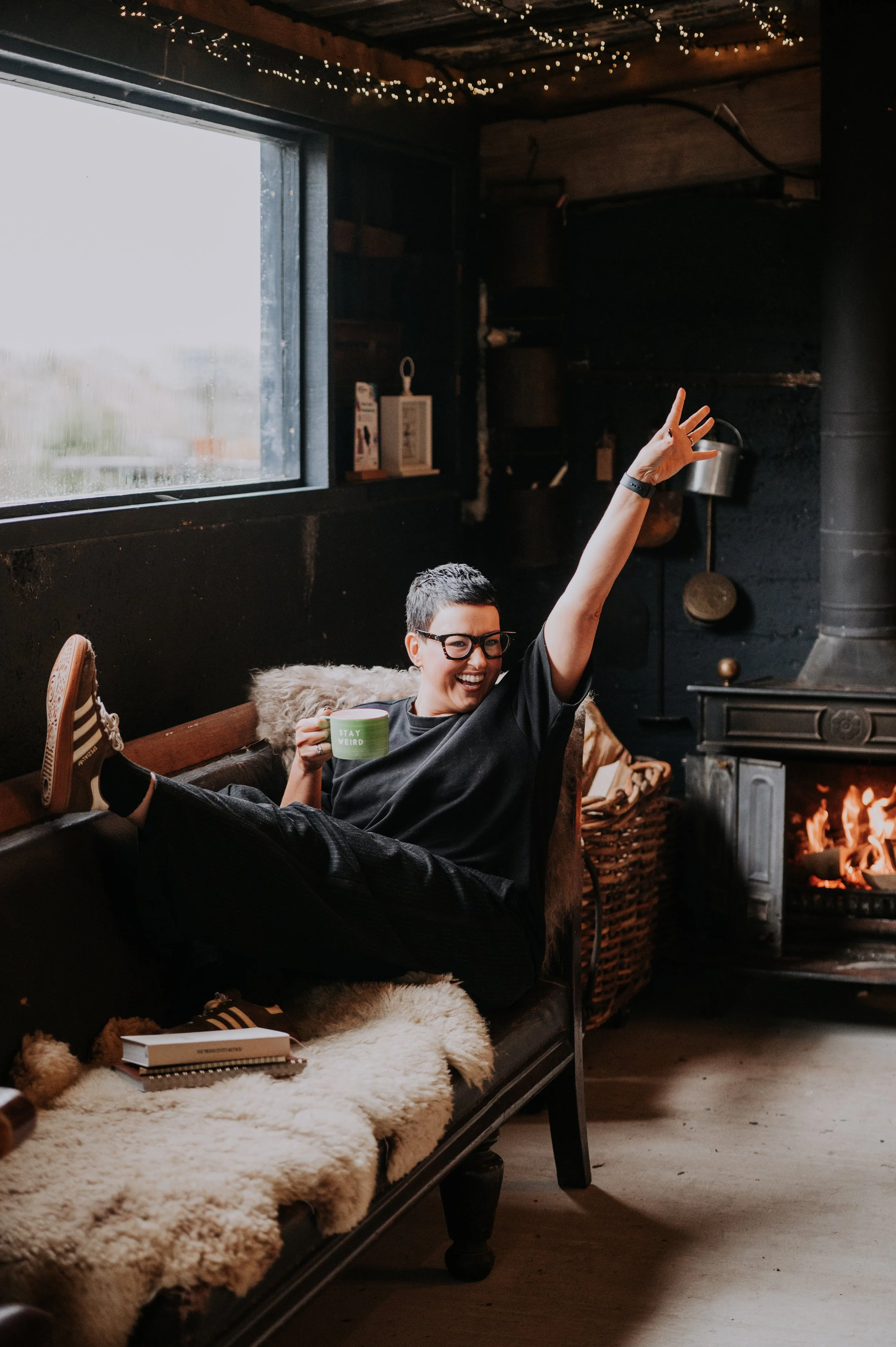 A person wearing glasses and a black shirt sitting on a black sofa, smiling and raising one arm, holding a green mug with 'STAY WEIRD' written on it, by a fireplace in a cozy, dimly lit room with string lights on the ceiling. There is a fluffy blanket, a book, and shoes on the sofa.