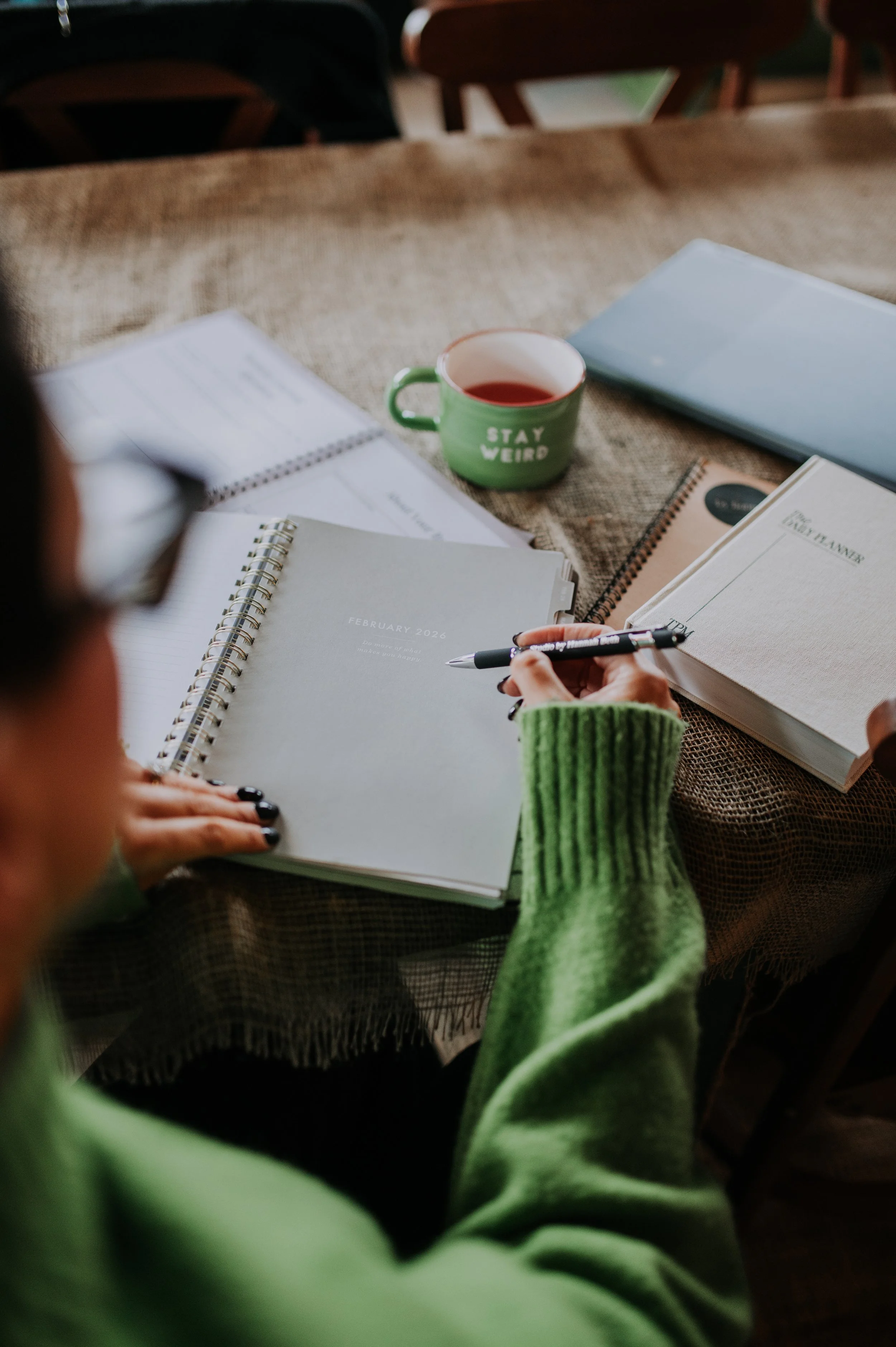 A person in a green sweater writes in a notebook on a table with many planners, a pen, a laptop, a mug that says 'Stay Weird,' and other planning materials. The setting appears to be a cozy indoor workspace or cafe.