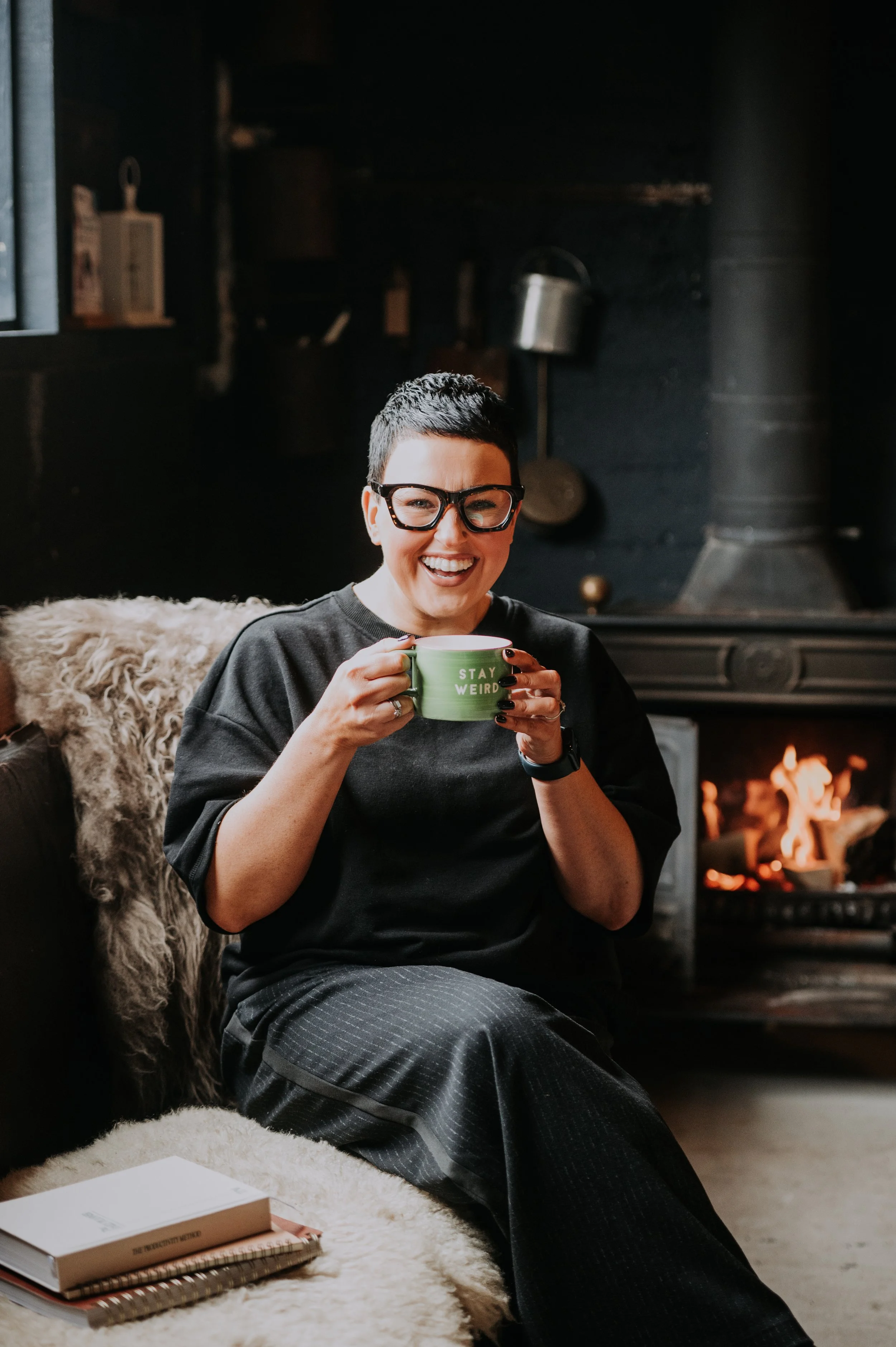 A woman with short dark hair and glasses, smiling and holding a green mug, sitting in a cozy living room with a fireplace, a furry blanket, and a book on a fluffy rug.