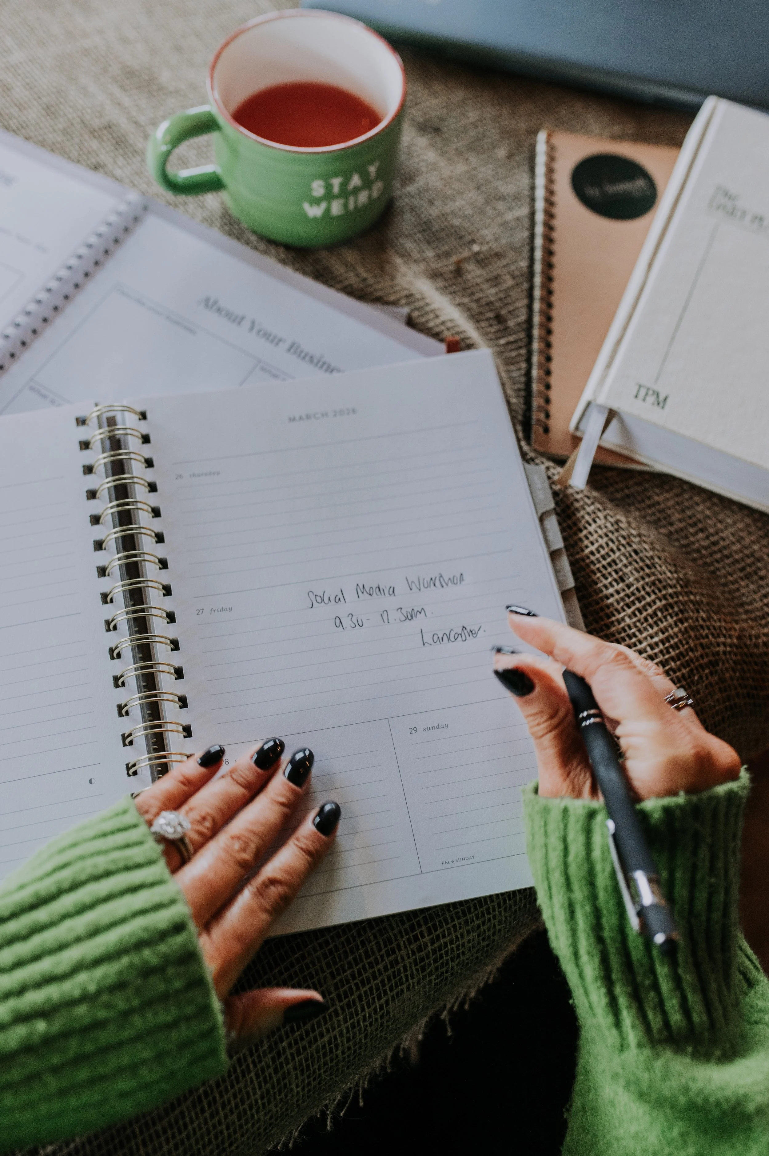 A person with black nail polish and rings is writing in a planner with a black pen. The planner is open to date March 2023, with a schedule for social media writing from 9:30 to 11:30 am. The person is wearing a bright green sweater. There are other notebooks, a green mug with red liquid, and a textured surface in the background.