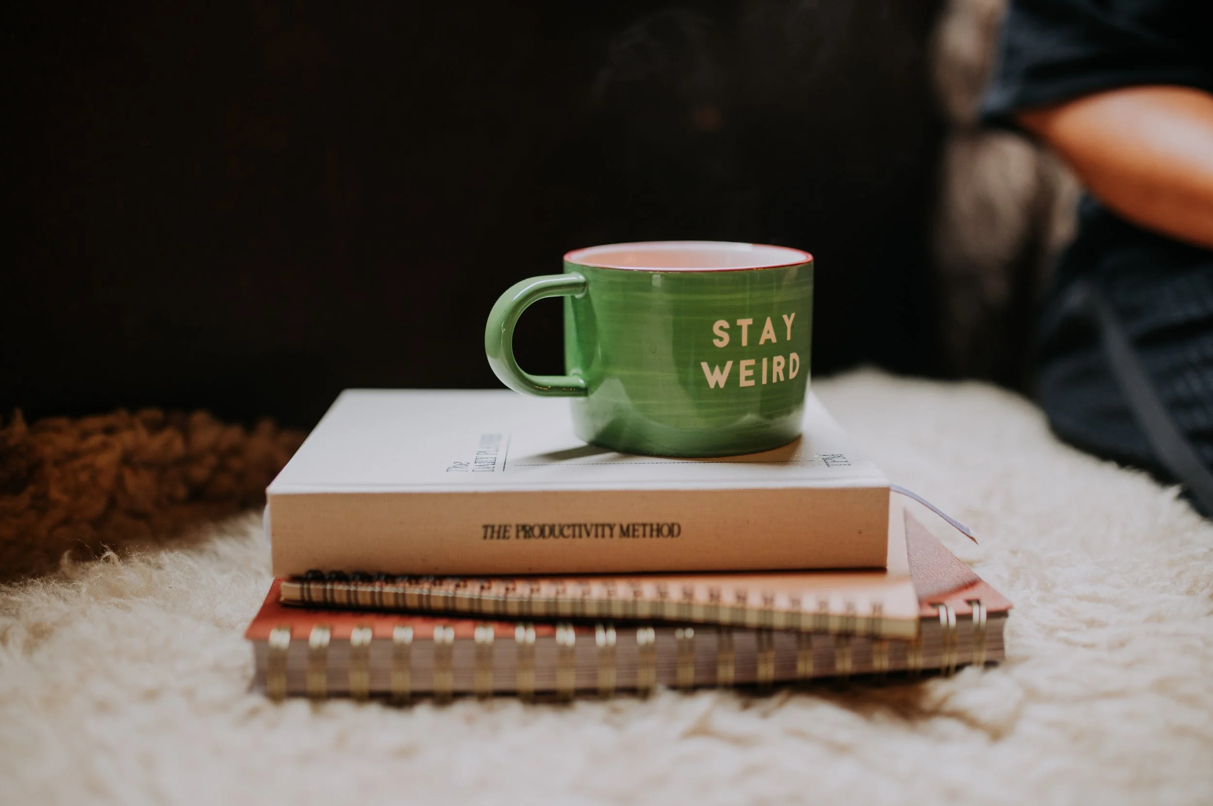 Green mug with the words 'STAY WEIRD' written on it, placed on top of a closed book titled 'The Productivity Method' and surrounded by additional notebooks and papers on a fluffy surface.