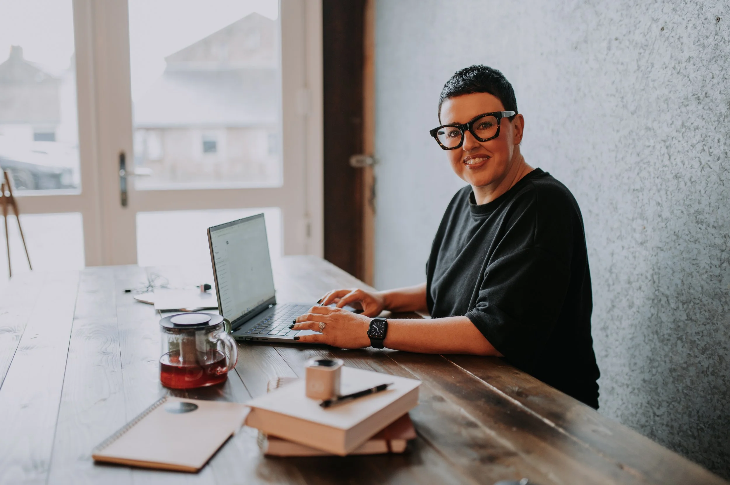 Woman with short dark hair and glasses sitting at a wooden table using a laptop. There are books, a notebook, a cup of tea, and other items on the table. A window and door are in the background.