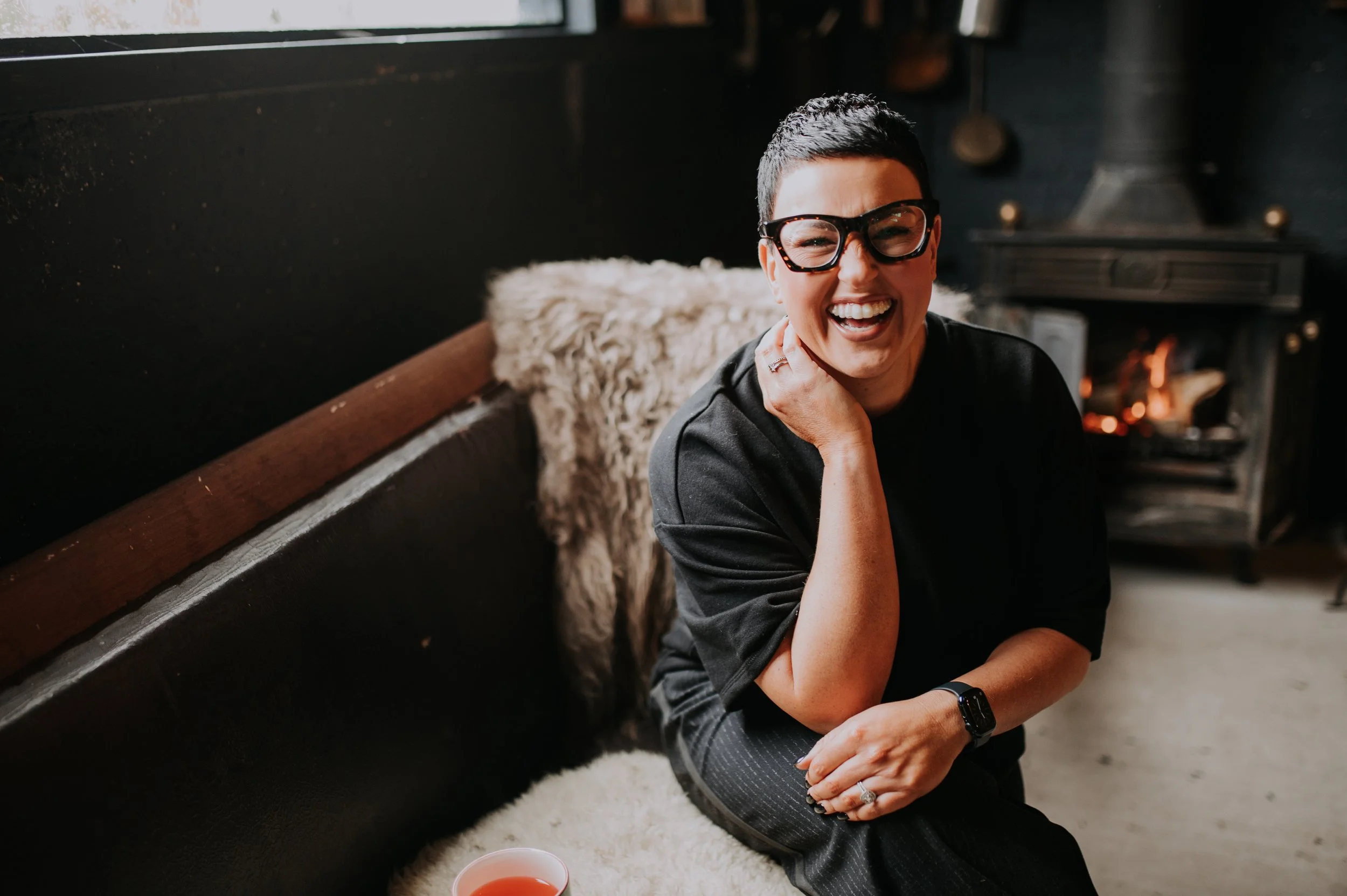 A woman with short dark hair, wearing glasses and a black shirt, smiling while sitting on the floor near a fireplace with her hand on her neck. A cup of tea or coffee is in front of her, and a cozy rug is underneath.