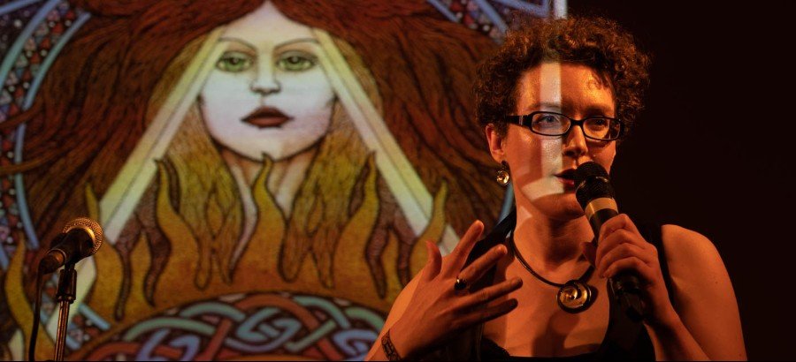 Photo of Sorcha Hegarty talking into a mic with one of the St Brigid Illuminations in the background.