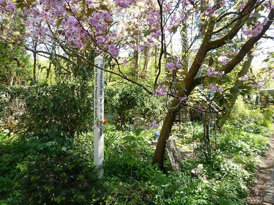 Photo pf the Peace Pole surrounded by greenery and a Cherry Blossom Tree.
