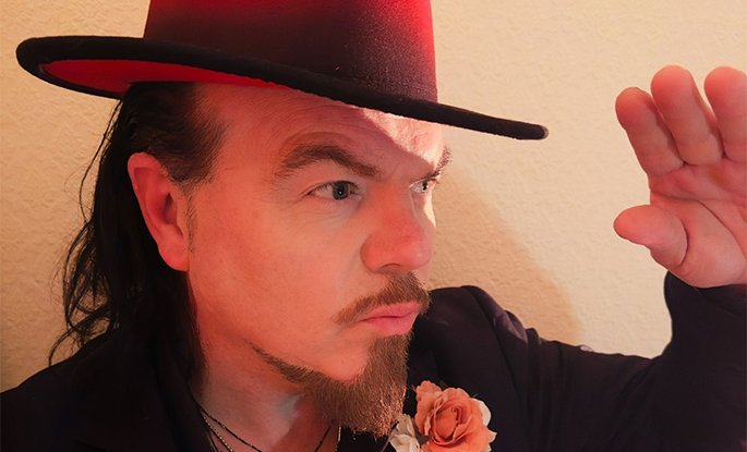 Close up photo of singer Jack Lukeman, wearing a red hat , black suit jacket and orange rose on his lapel.