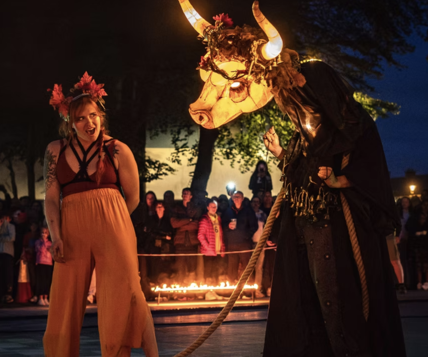 Photo of storyteller talking to a lit up larger than life bull puppet, onlookers are dressed cosy while candles create a firey atmosphere