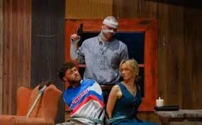 Photo from the play The burned Mana man hold a gun a man and woman are tied up together in a sitting room.