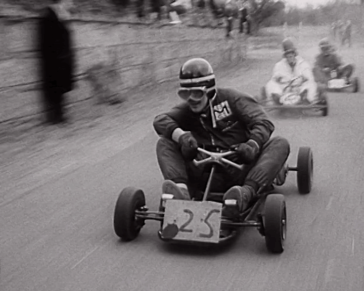 Black and White Photo of a man in full racing gear with a helmet , gloves and goggles driving a go Kart image is from around the 1970’s