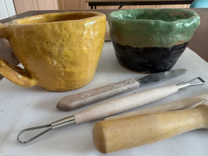 CLose up photo of a custard yello handmade tea cup and a pale green and navysugar bowl, with pottery tools in the foreground , background with tables