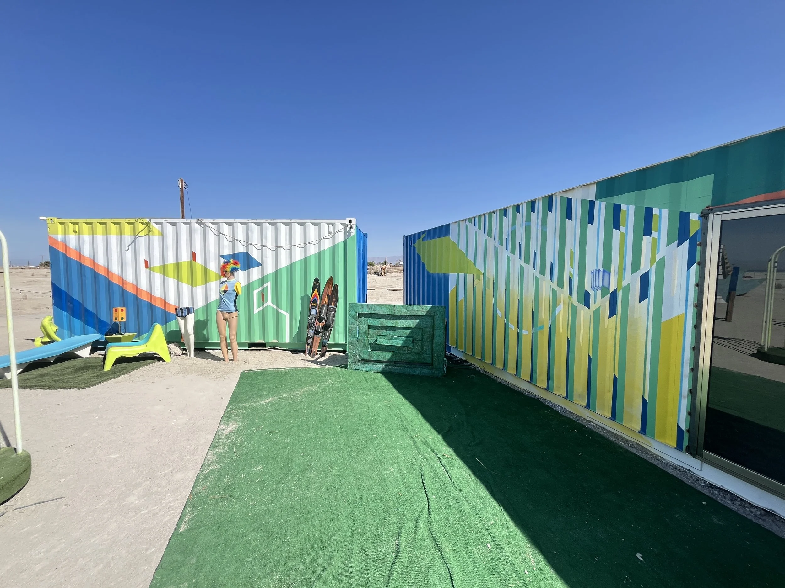 Colorful beachside art installation with geometric painted shipping containers, a mannequin with colorful sunglasses, surfboards, and artificial grass, under a clear blue sky.