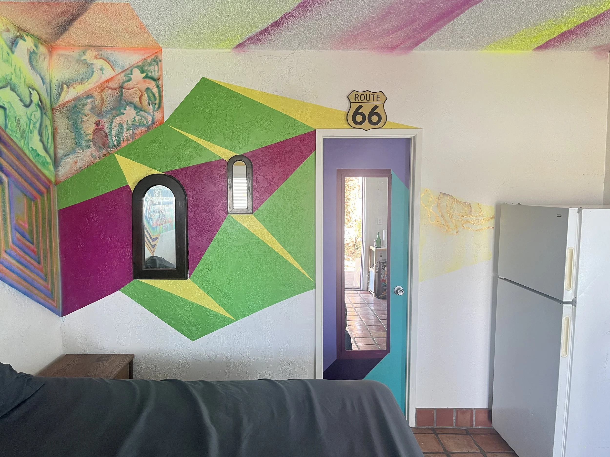 Interior wall with colorful geometric mural, small arched and rectangular mirrors, Route 66 sign, doorway with purple and blue walls leading outside, refrigerator, and part of a sofa in the foreground.