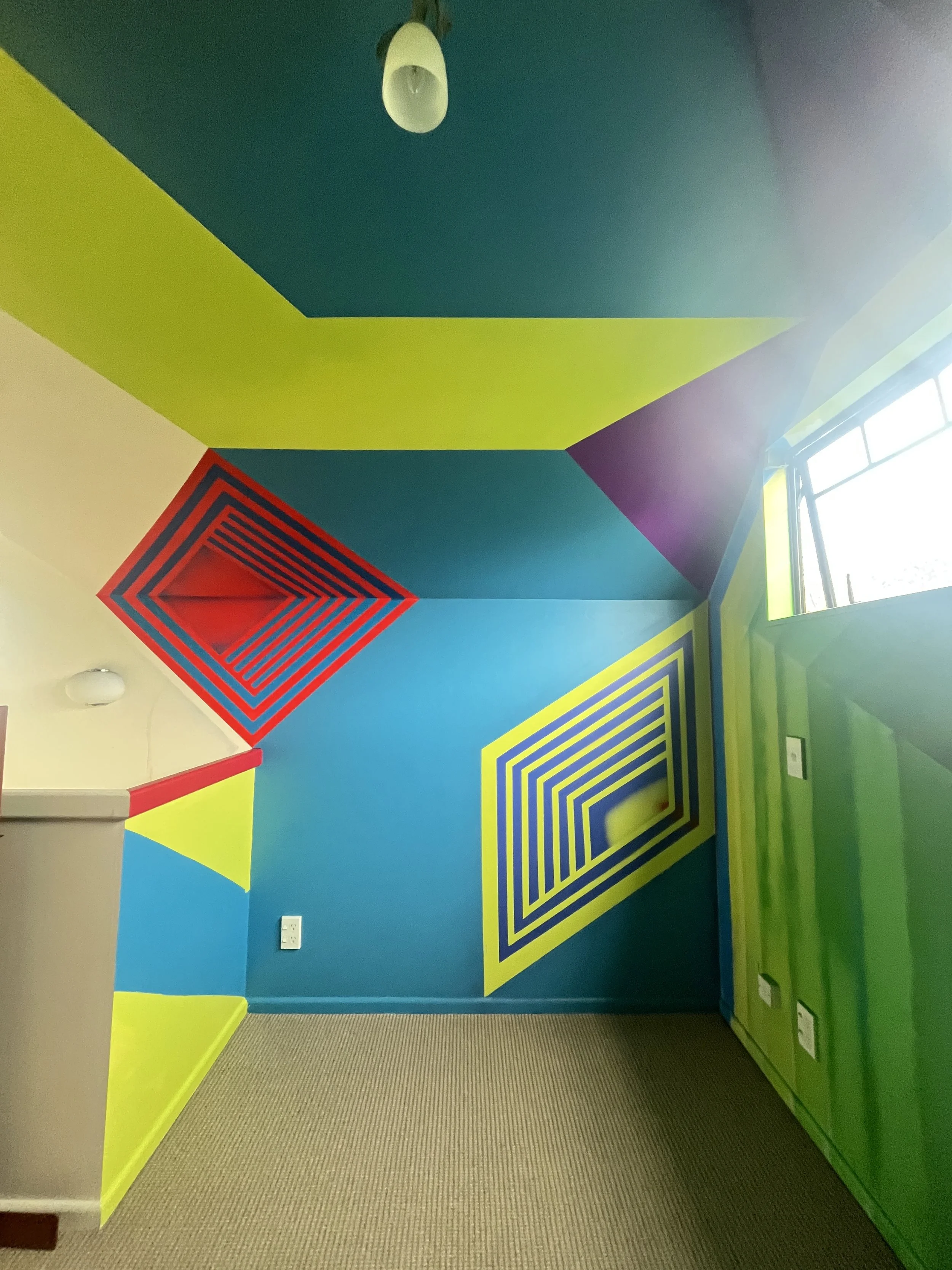 Colorful geometric wall mural with concentric squares and triangles in red, yellow, blue, purple, and green in a room with a window.