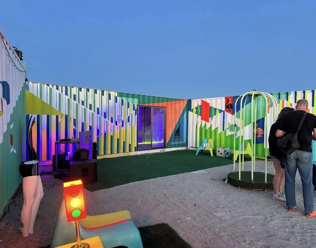 A colorful outdoor play area with striped painted walls, a small slide shaped like traffic lights, a miniature table and chairs, and inflatable fun elements under a clear blue sky.