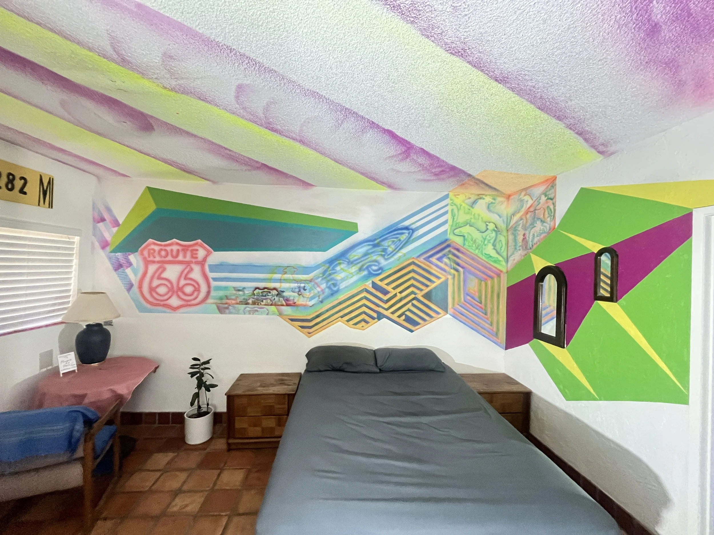 A bedroom with a colorful mural on the wall, featuring a Route 66 sign, abstract geometric patterns, and vibrant colors. The room contains a bed with gray bedding, wooden nightstands, a small potted plant, a lamp, and additional furniture.