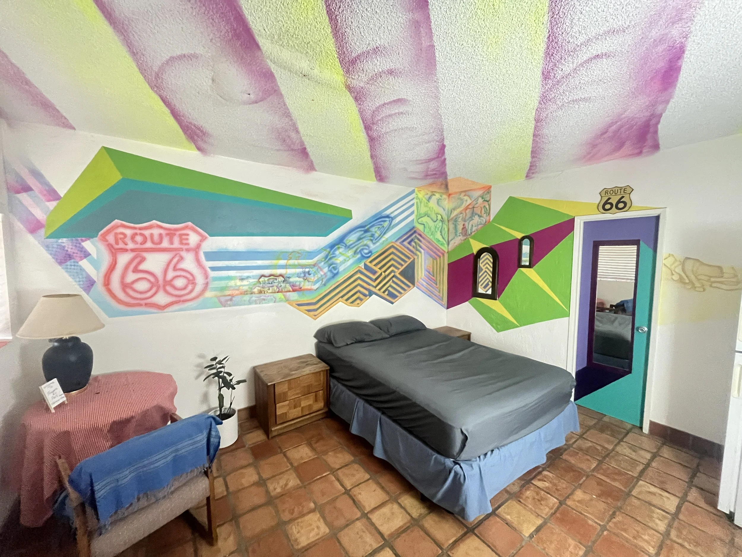A bedroom with colorful graffiti-style wall art, a bed with dark sheets, a bedside table, a lamp, a small chair with a blue cover, and a small potted plant. The ceiling has pink, purple, and green abstract designs, and the door has a multicolor geometric pattern with a Route 66 sign above it.
