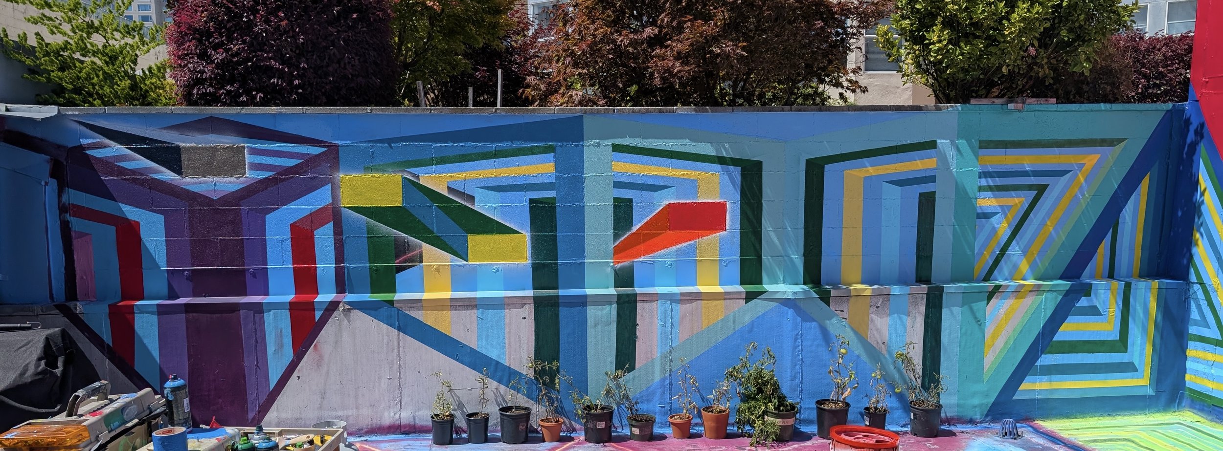 Colorful geometric mural painted on a wall with abstract shapes and patterns in shades of blue, green, yellow, red, and purple. There are potted plants at the bottom and trees in the background.