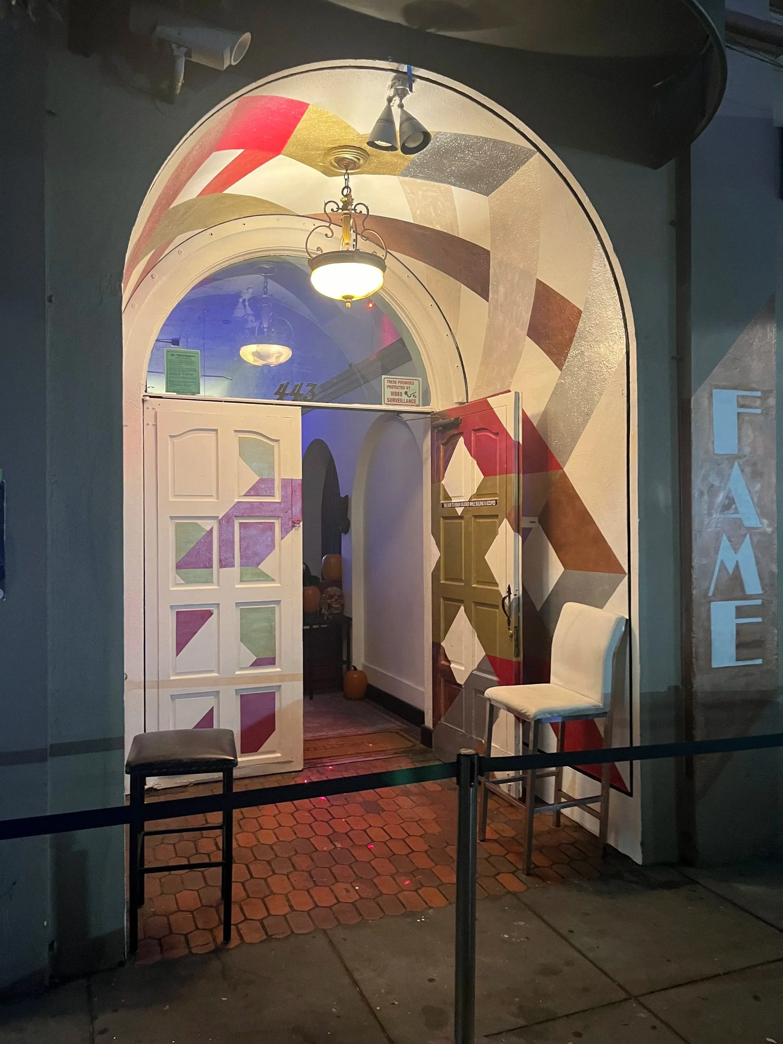 The image shows an outside view of a building entrance with a large open door leading into a room with a colorful geometric mural on the wall. There are chairs outside, a small step, and a barrier rope in front of the entrance. The interior visible has pumpkins and a cluttered side table, suggesting a festive or welcoming setting.
