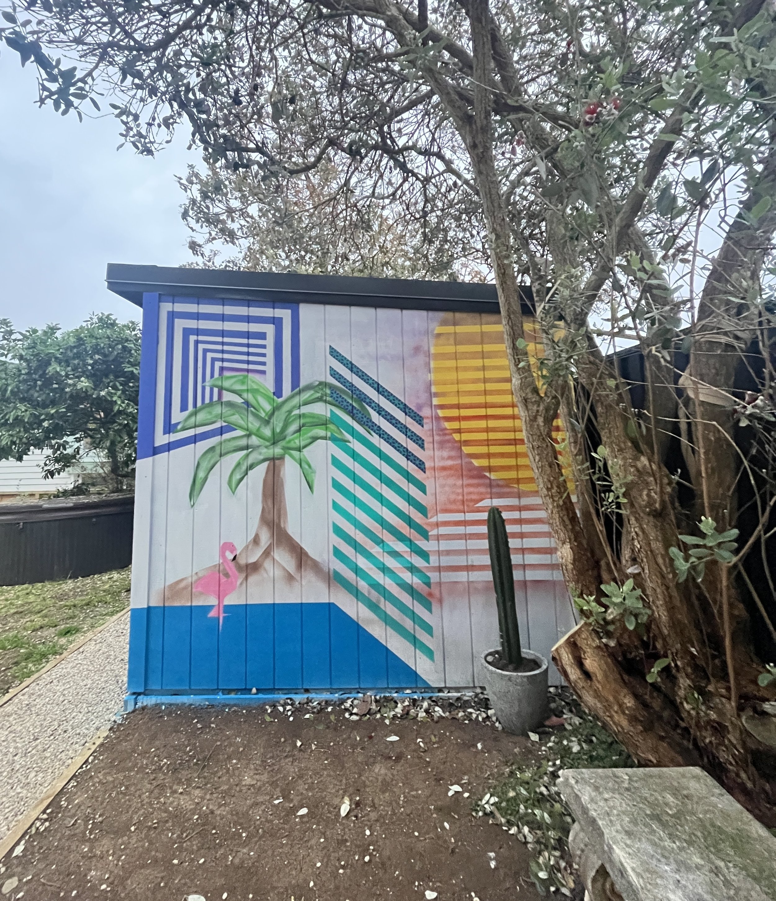 Colorful mural featuring a green plant, pink flamingo, stylized orange sun, and geometric patterns on a wooden wall next to a tree and cactus.