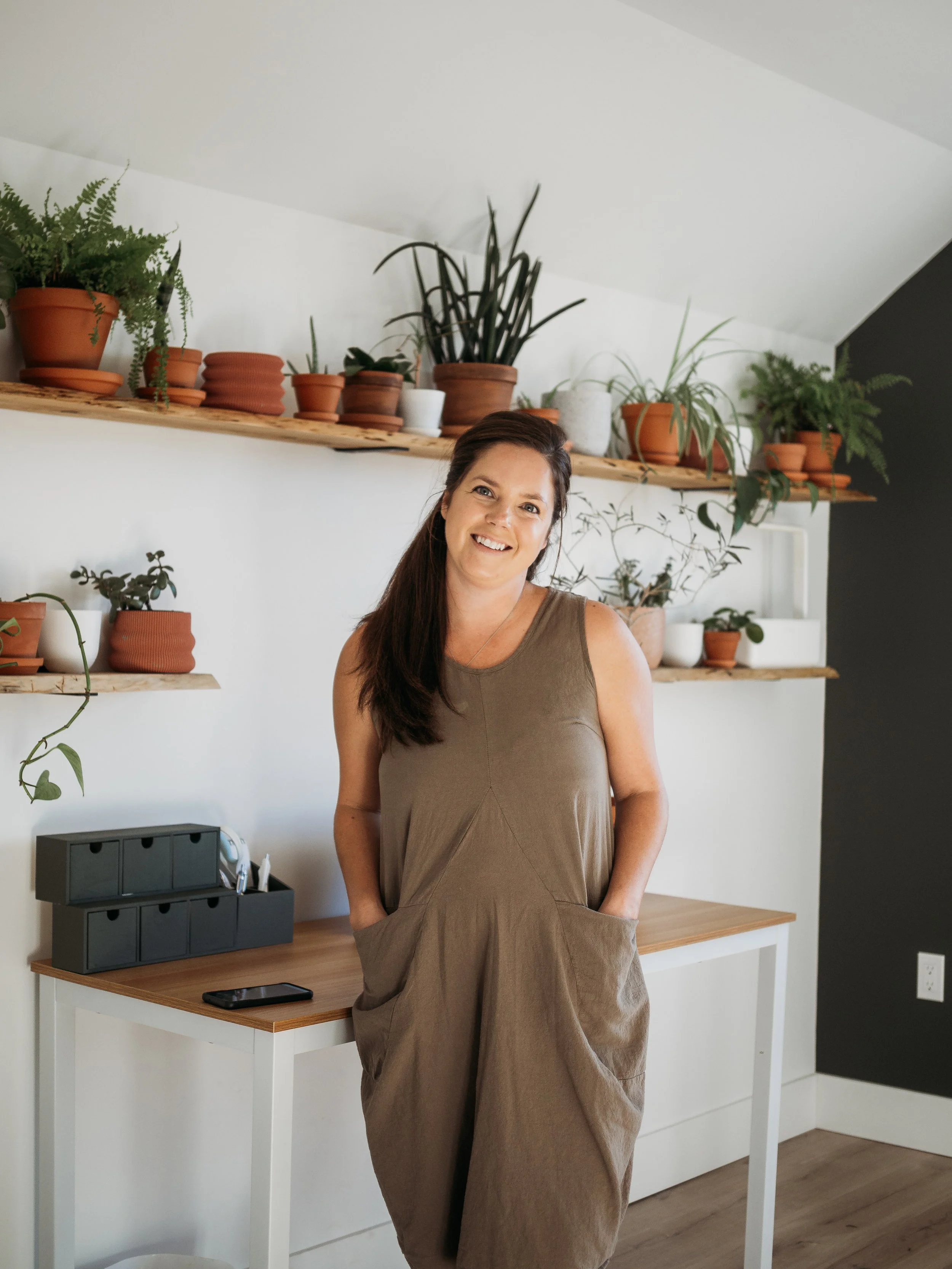 A woman with long dark hair smiling, wearing a brown sleeveless dress, standing in front of a white wall with two wooden shelves filled with various potted plants.