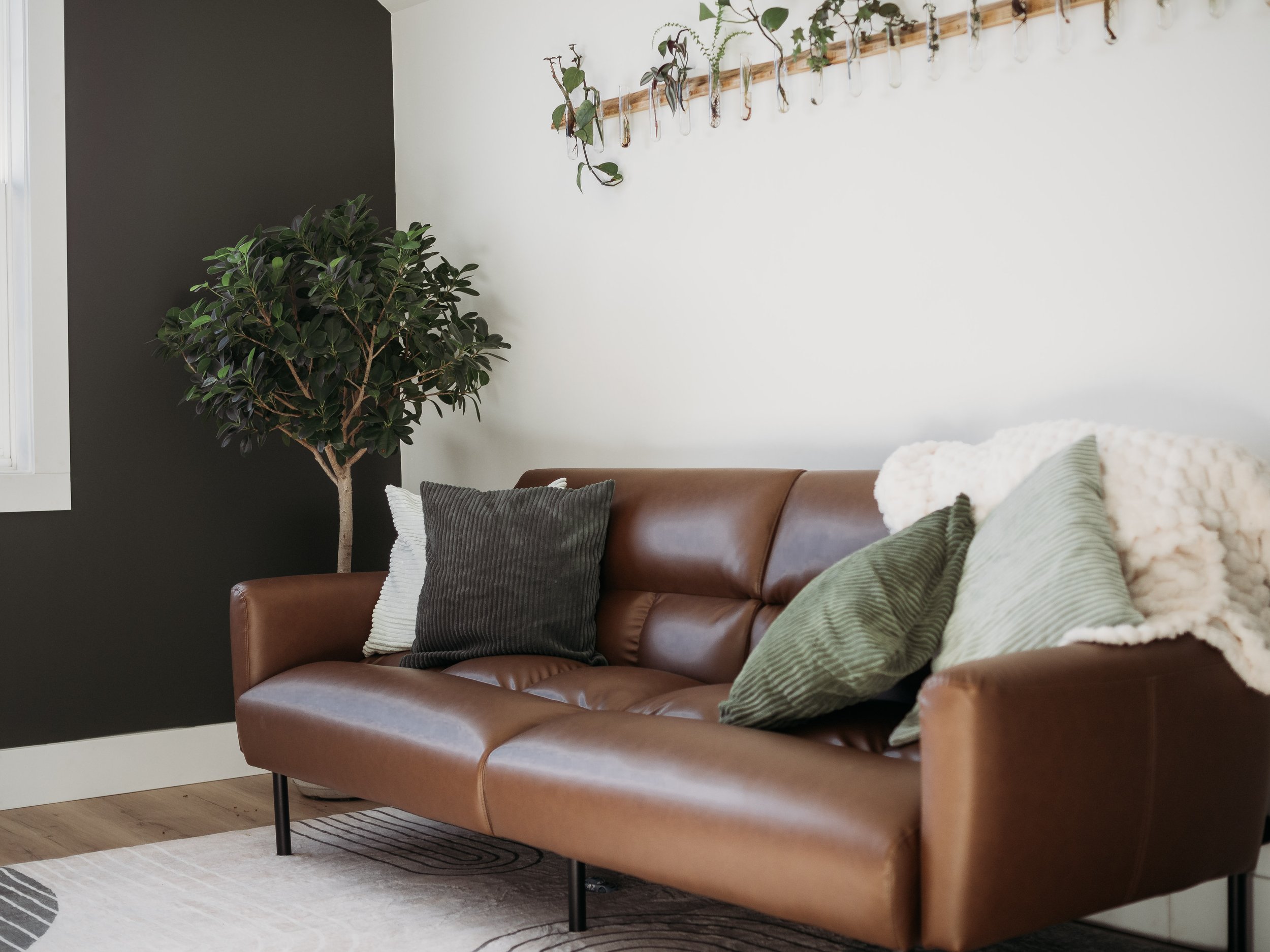 Living room with a brown leather sofa, green and white pillows, a fluffy white blanket, a large potted plant, and a wall with a wooden coat rack with hanging plants.