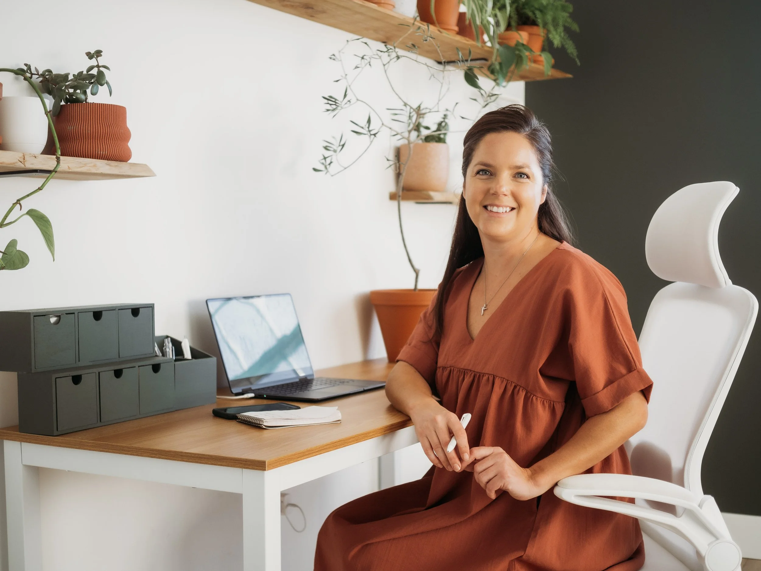 A woman sitting at a desk with a laptop, notebooks, and a desk organizer in a modern, plant-decorated workspace, smiling at the camera.