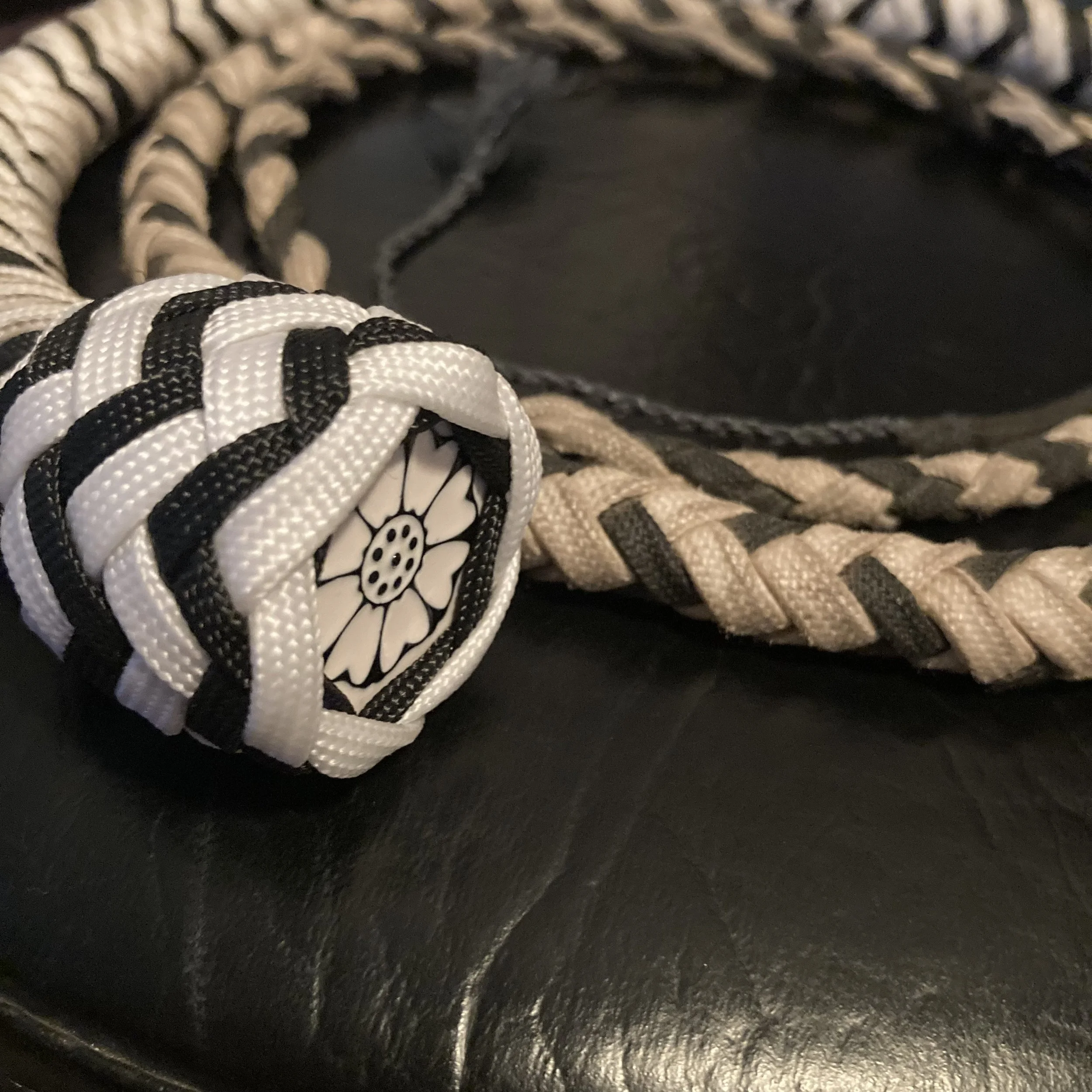 A close-up of a decorative rope knot with a black and white flower design on the end, resting on a black leather surface.