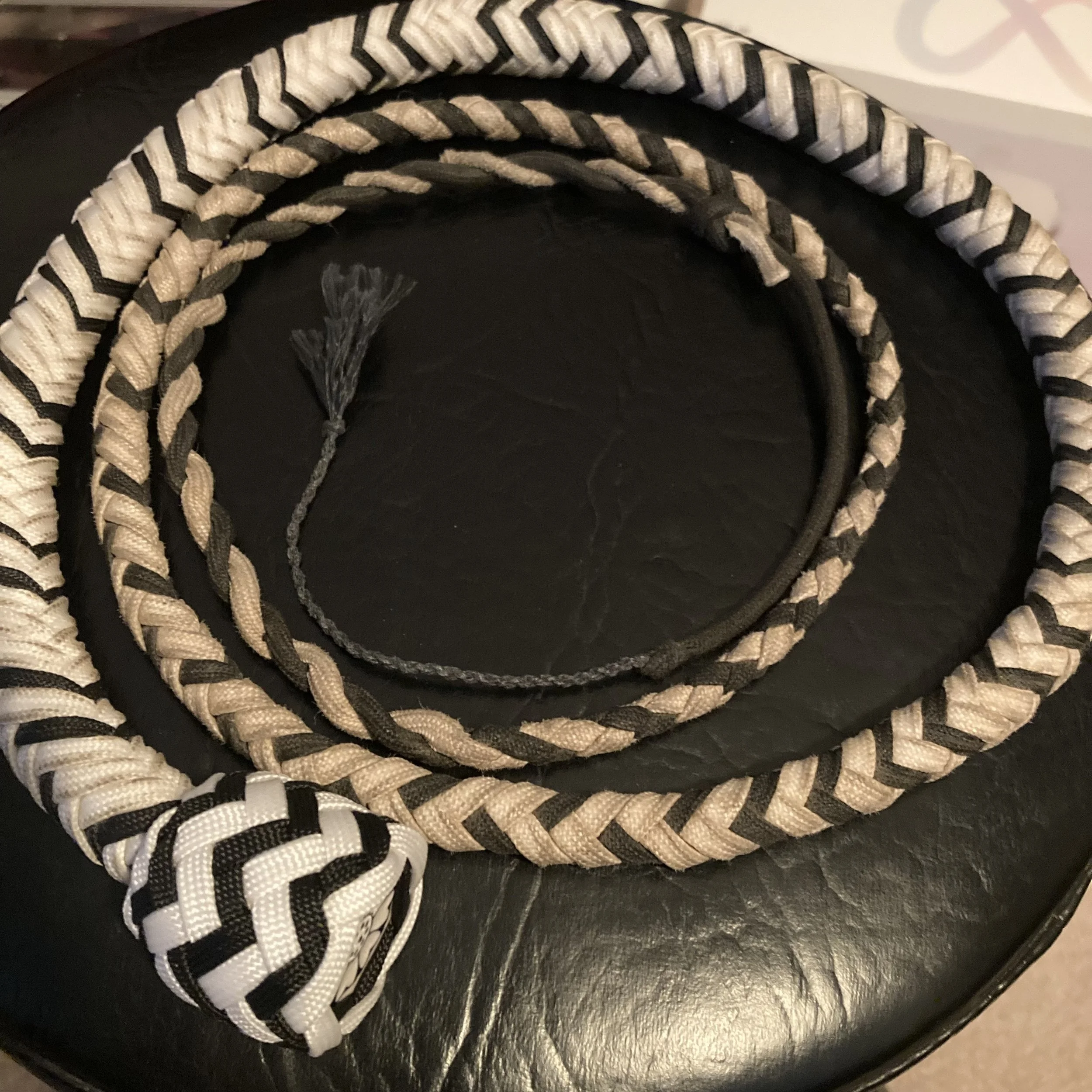 Three braided leash or rope leads in black, beige, and black-and-beige, with a knotted handle and tassel on a black leather surface.