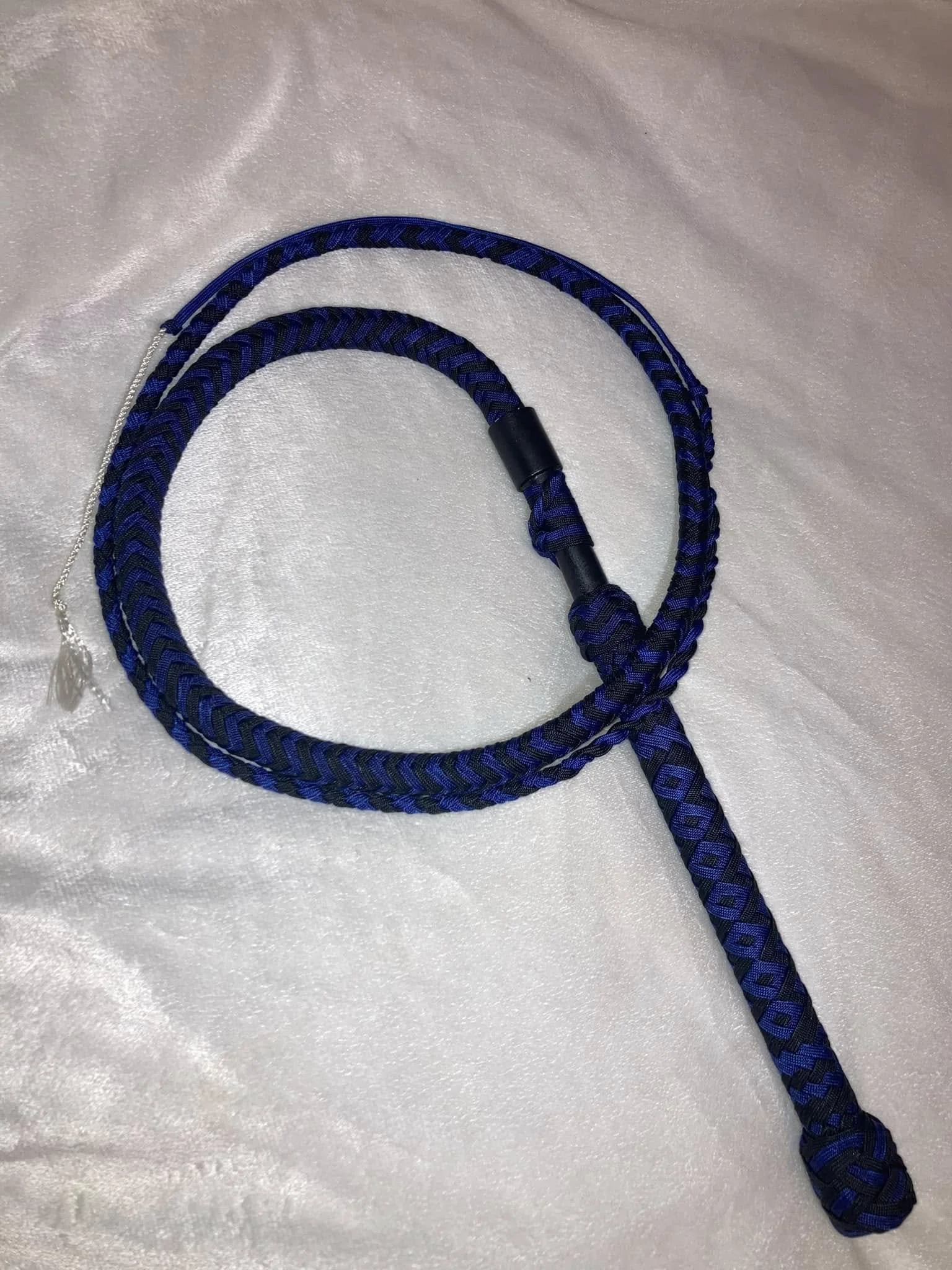 A blue and black braided rope with a loop and a small metal tassel at one end, lying on a shiny, light-colored fabric surface.