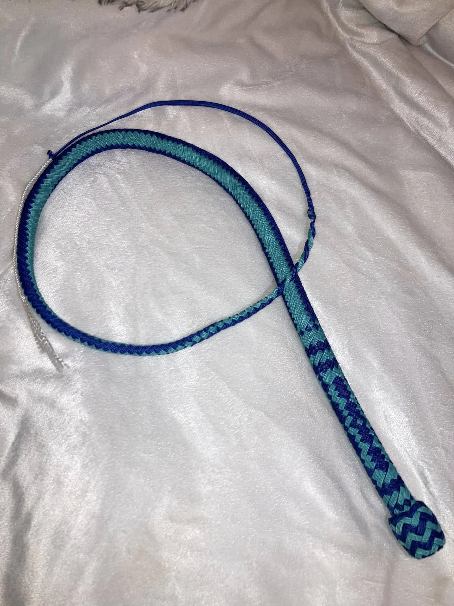A blue and turquoise braided whip with a loop handle, resting on a white satin fabric background.