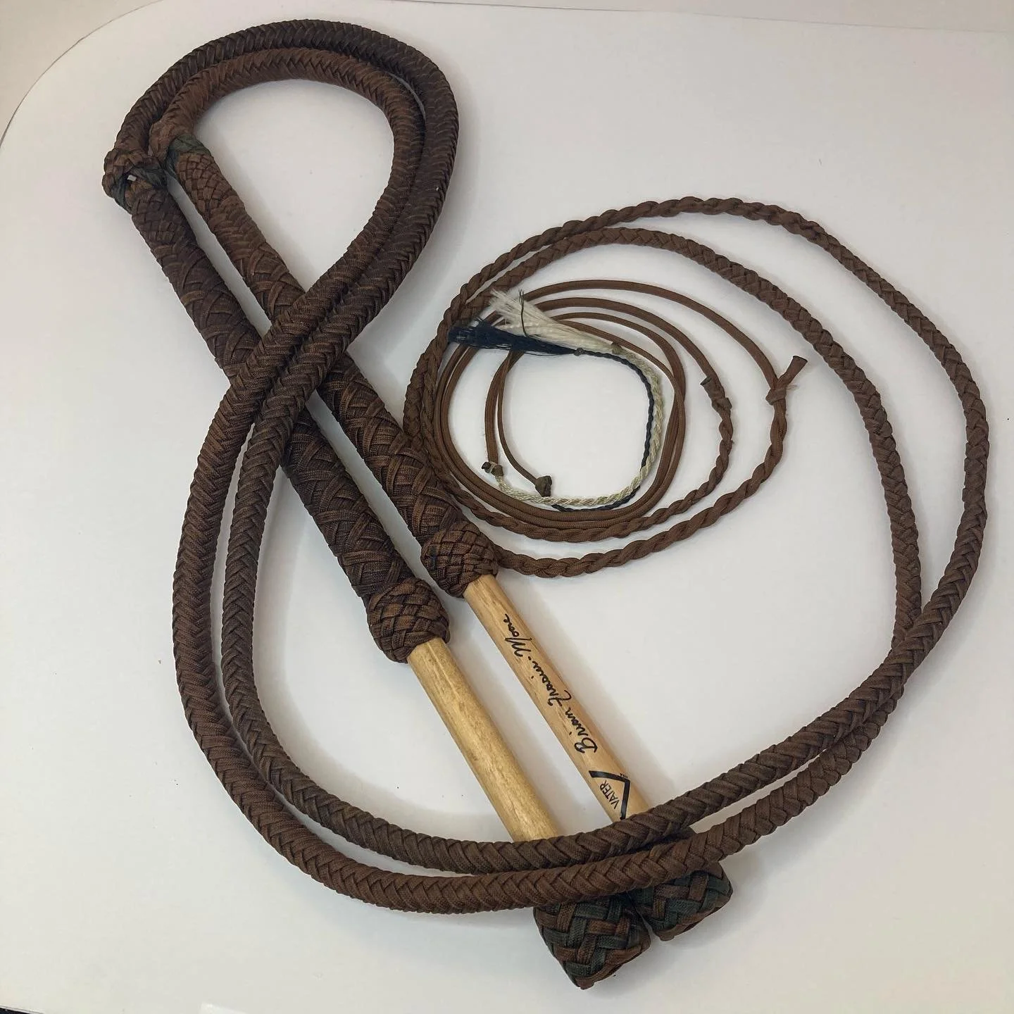 A brown braided whip with a wooden handle and a smaller brown braided whip, along with several thin, brown braided cords on a white surface.