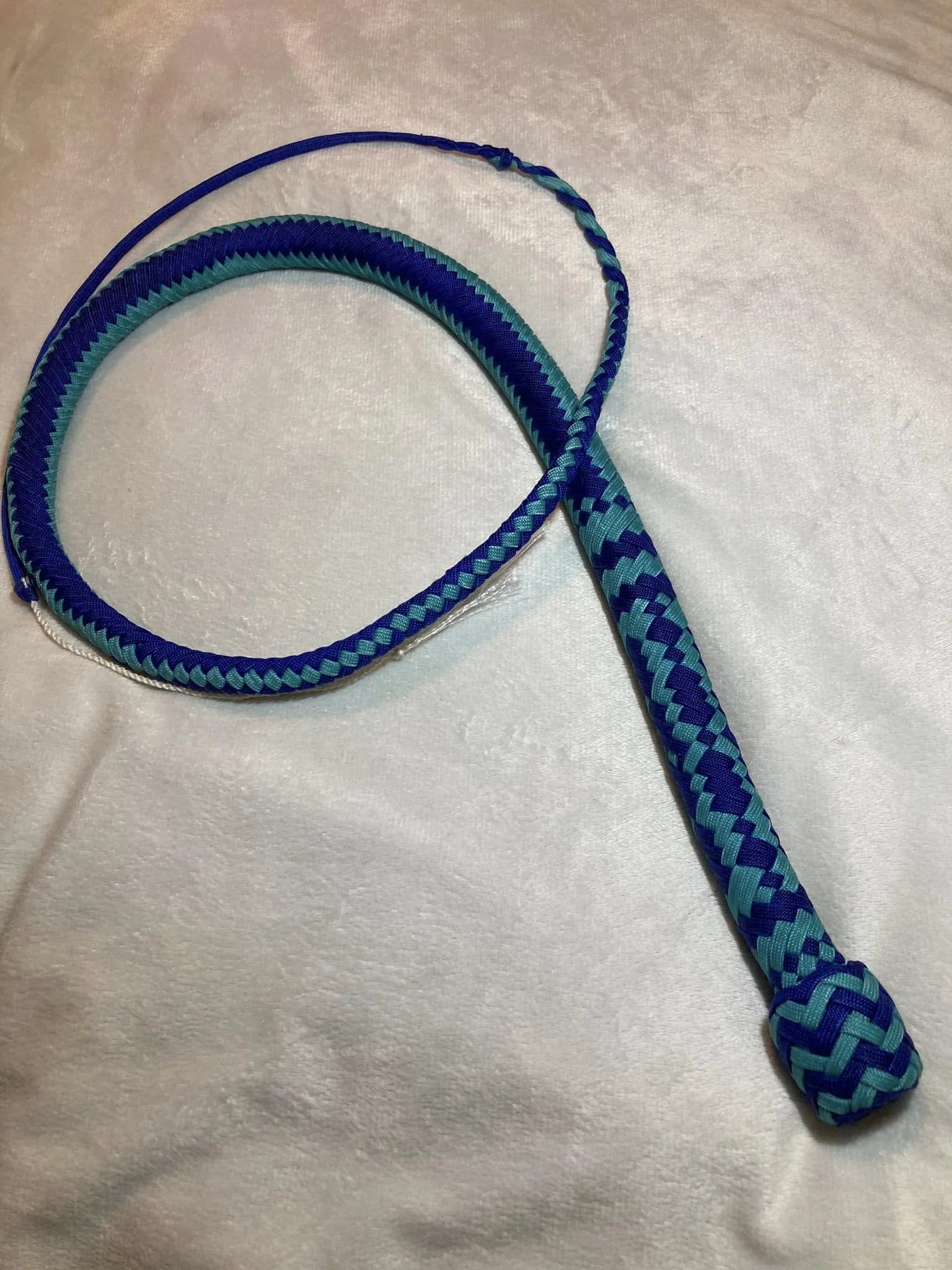 A braid tennis at of blue and turquoise in a diagonal pattern, with a black loop at one end, laying on a white fabric surface.