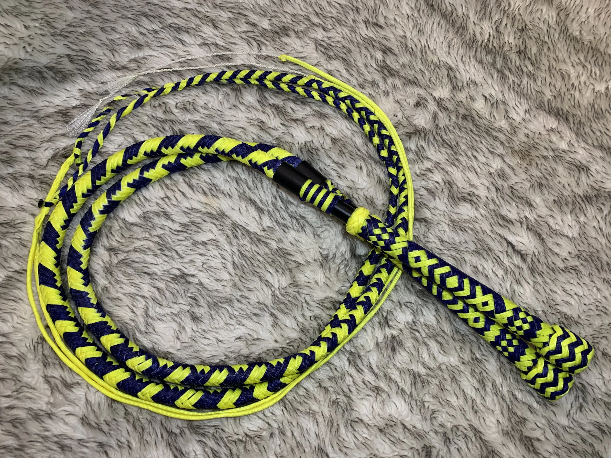 A skipping rope with a yellow, black, and navy blue braided design, lying on a beige fuzzy surface.