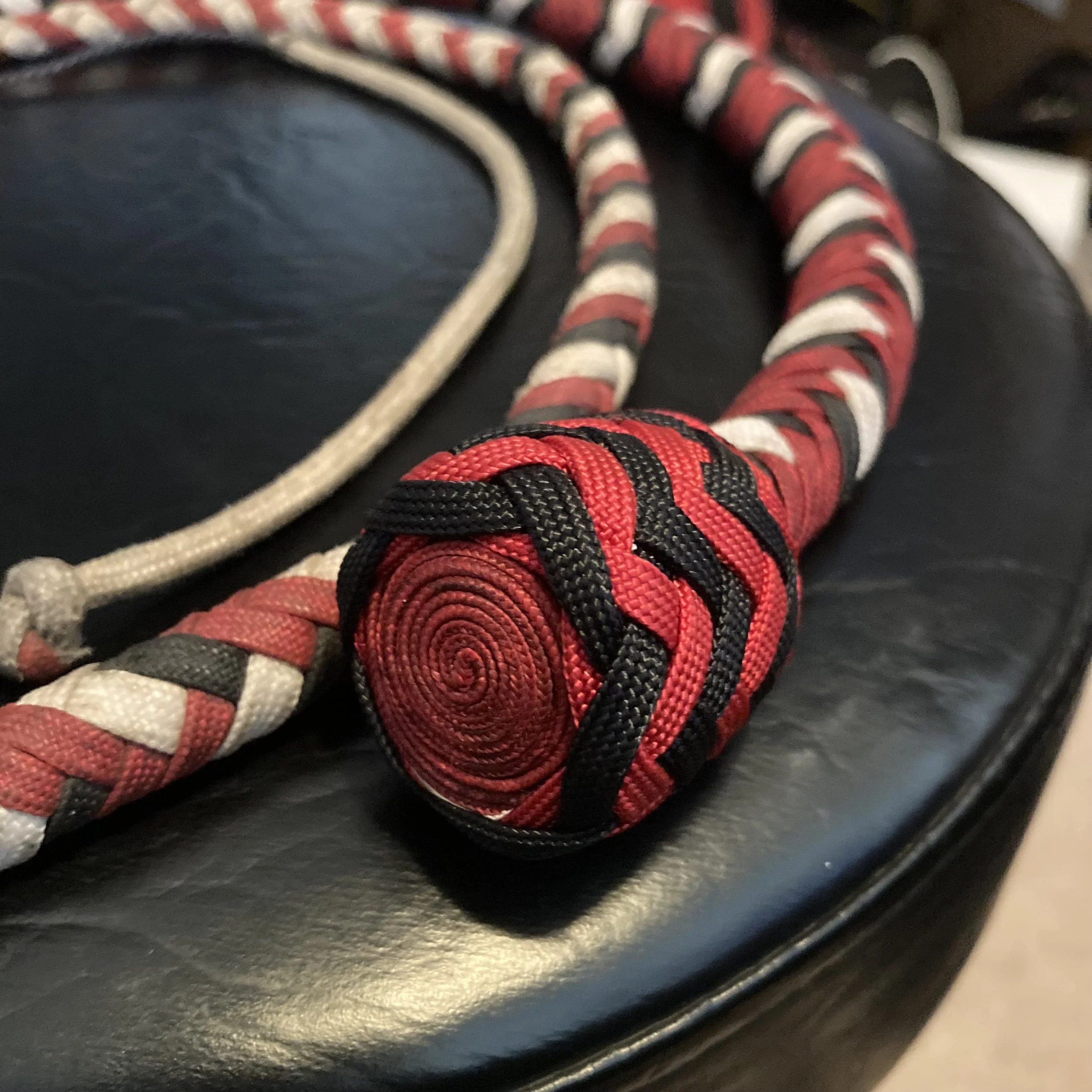 Close-up of a red, black, and white woven fabric whip or braided cord with a tightly wound handle resting on a black leather surface.