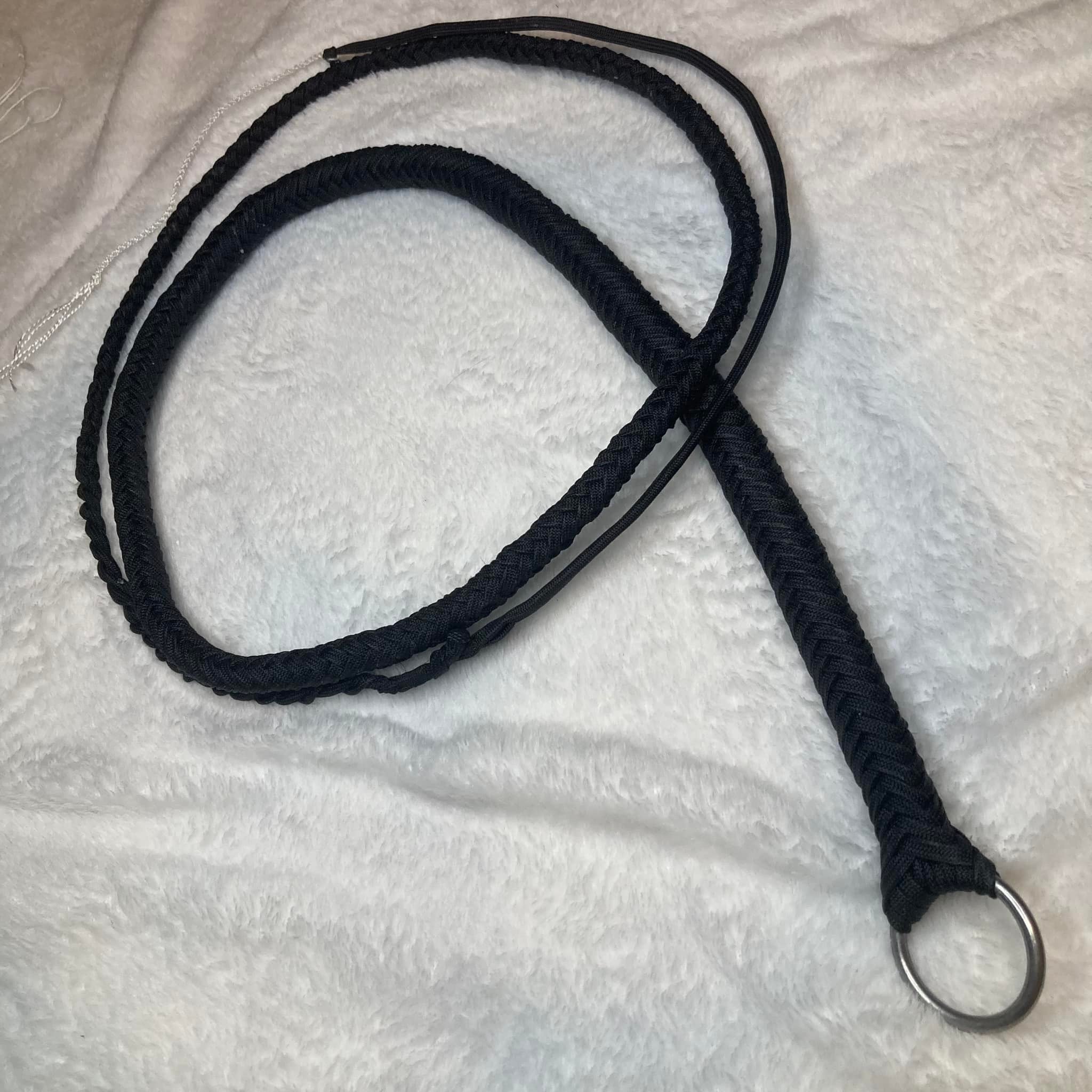 A black braided lanyard with a metal ring attached at one end, lying on a white textured fabric surface.
