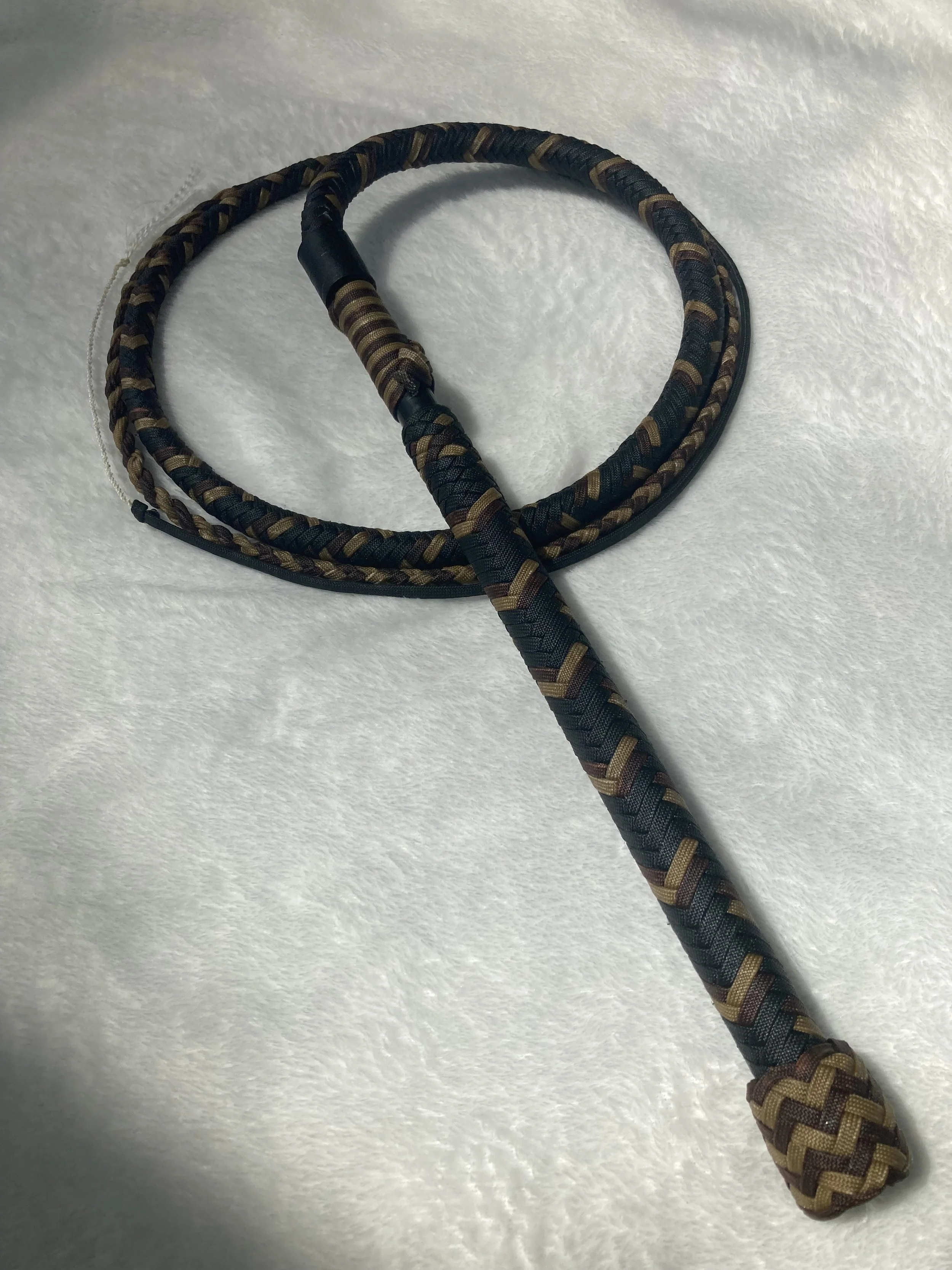 A patterned woven braided whip or switch with a handle, placed on a white textured surface.